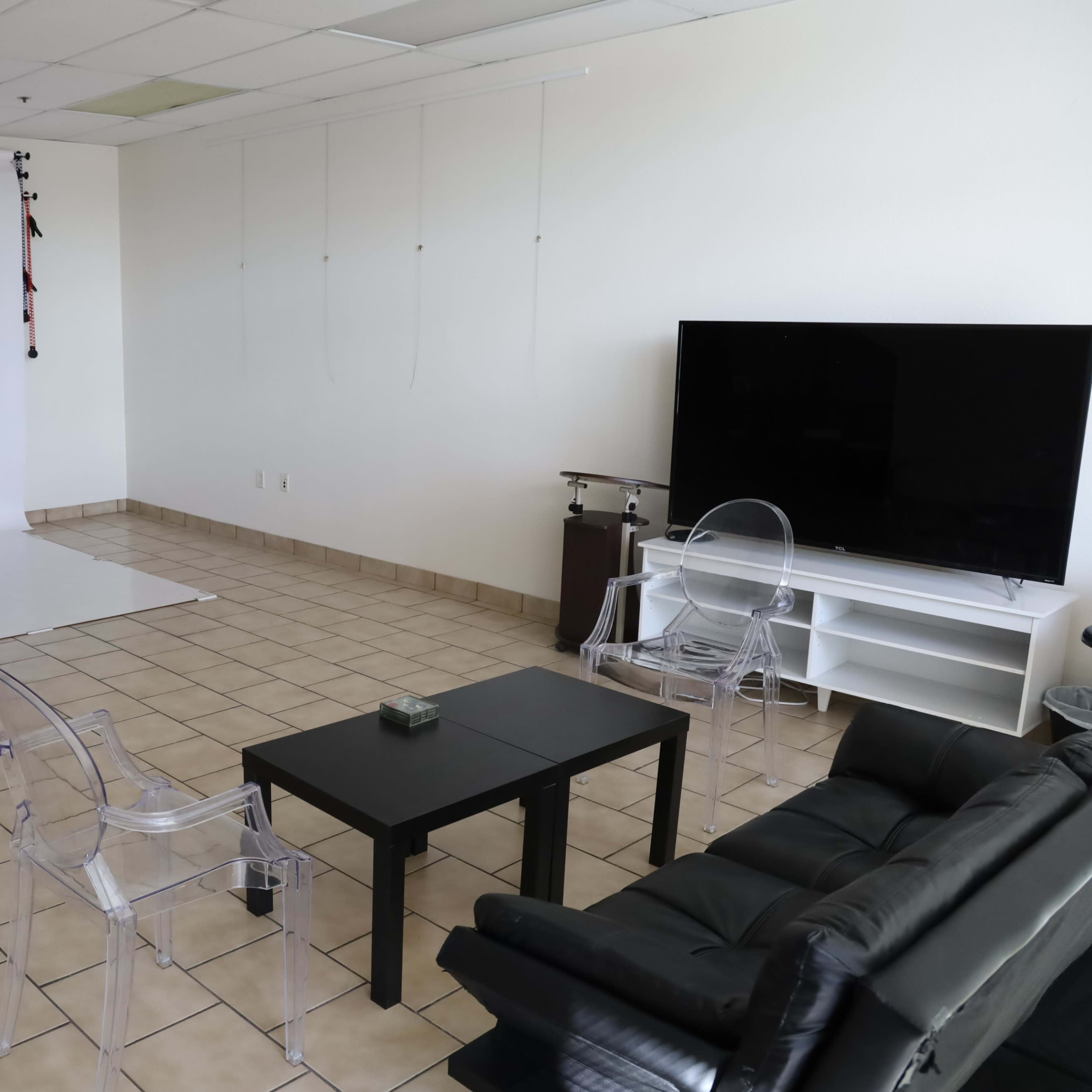 A minimalistic studio space with a plain white backdrop, a black table, transparent chairs, and a large television.