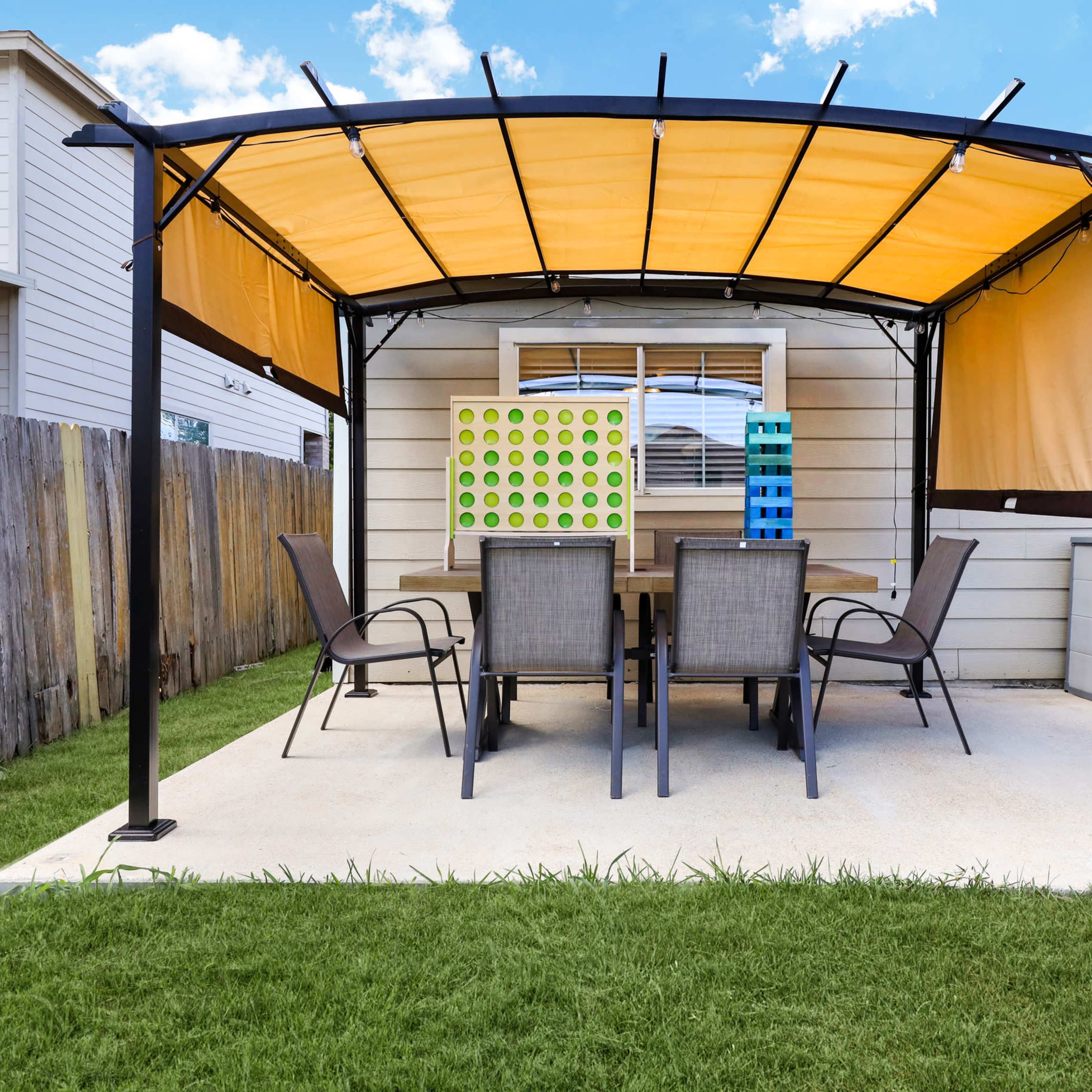 A patio with an orange canopy covers a table and six chairs in a backyard, with a grass lawn and a nearby shed.