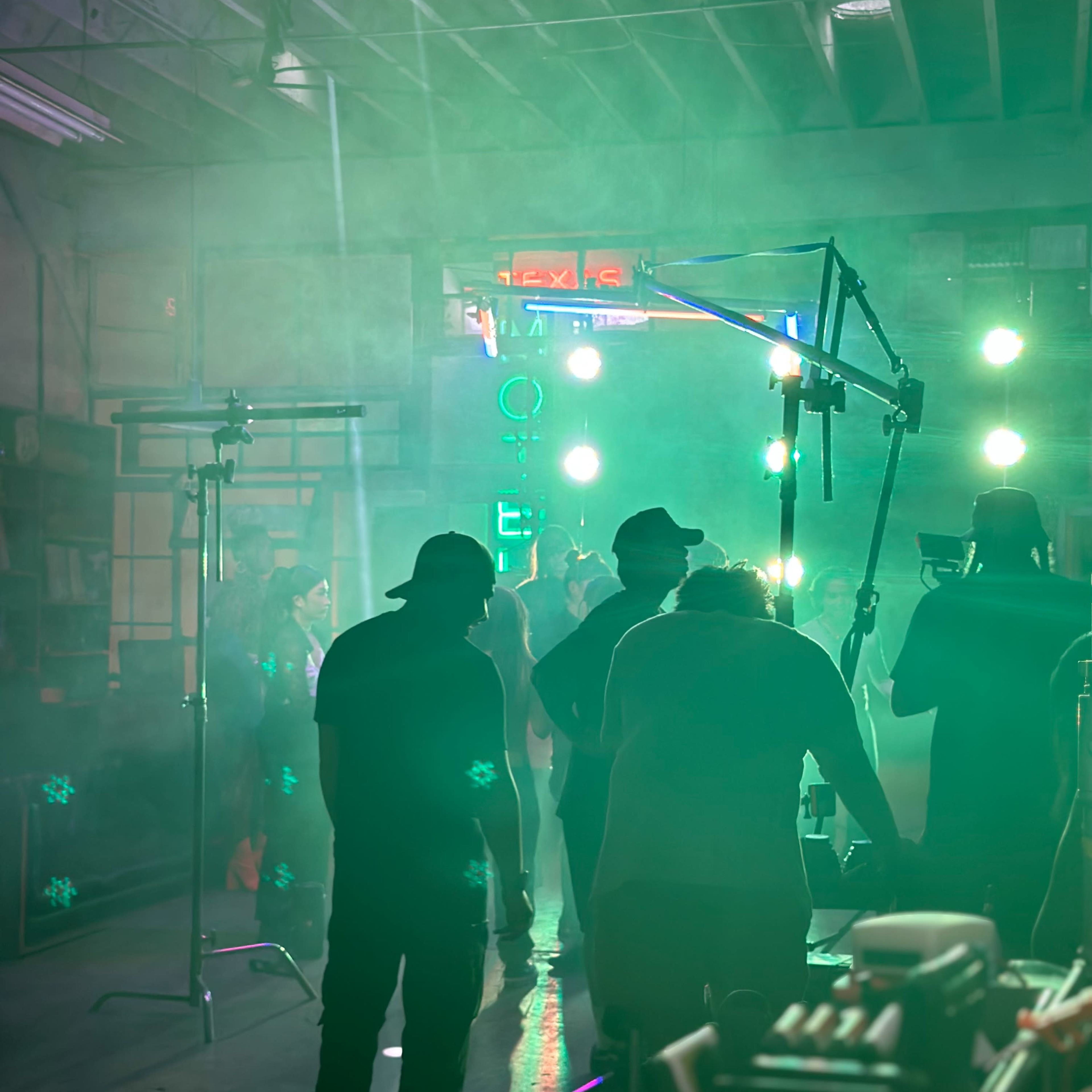 A group of people are silhouetted against green lighting in a dimly lit industrial space during a video shoot.