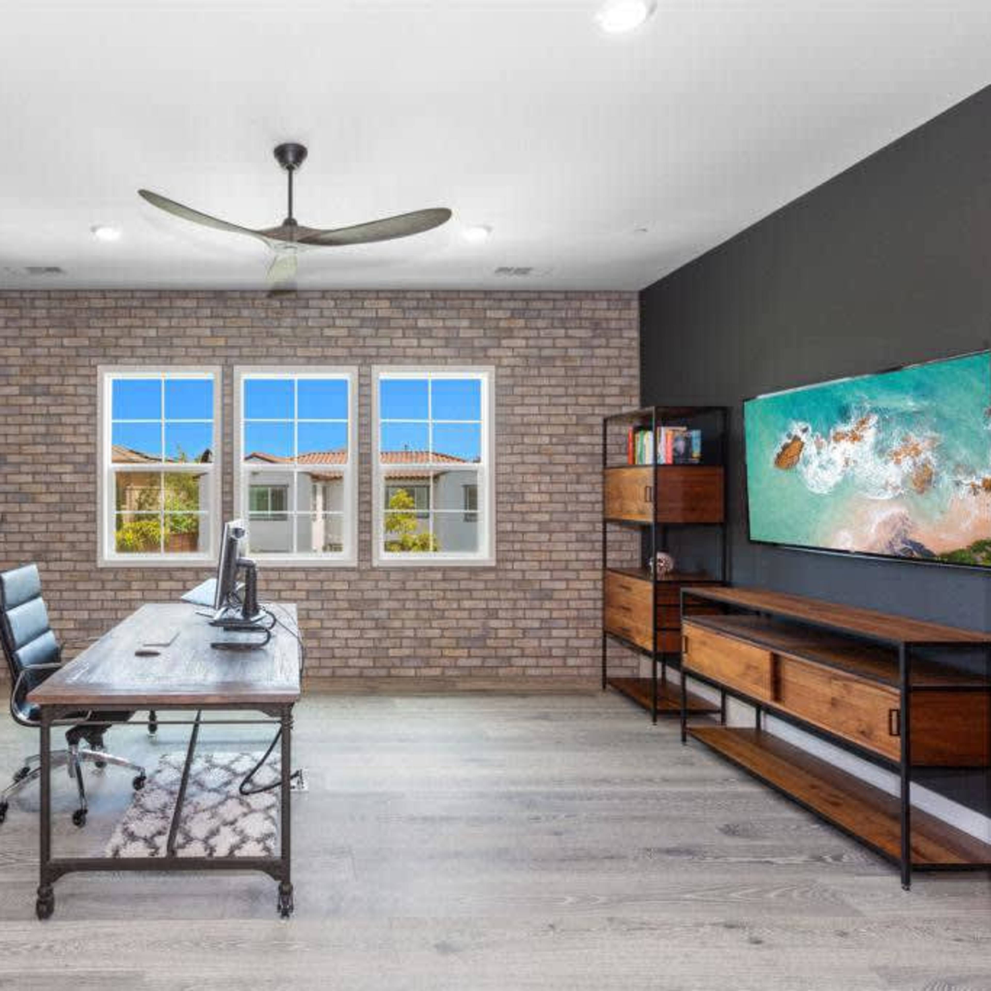 A modern office space with a brick accent wall, a wooden desk, a black chair, and large windows that provide natural light.