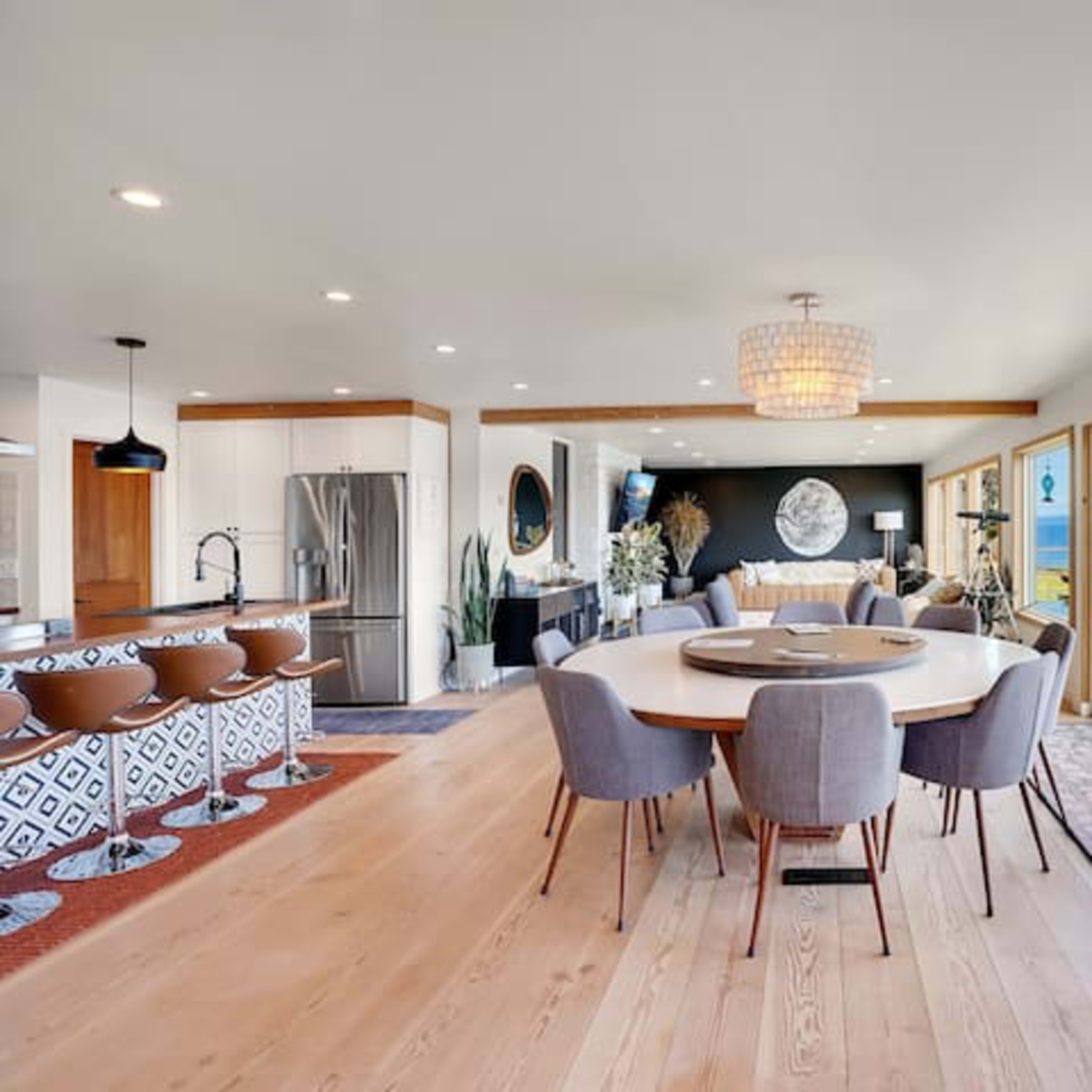 A bright, open-concept dining and living area with large windows overlooking a water view, featuring a round dining table surrounded by gray chairs and a modern kitchen.