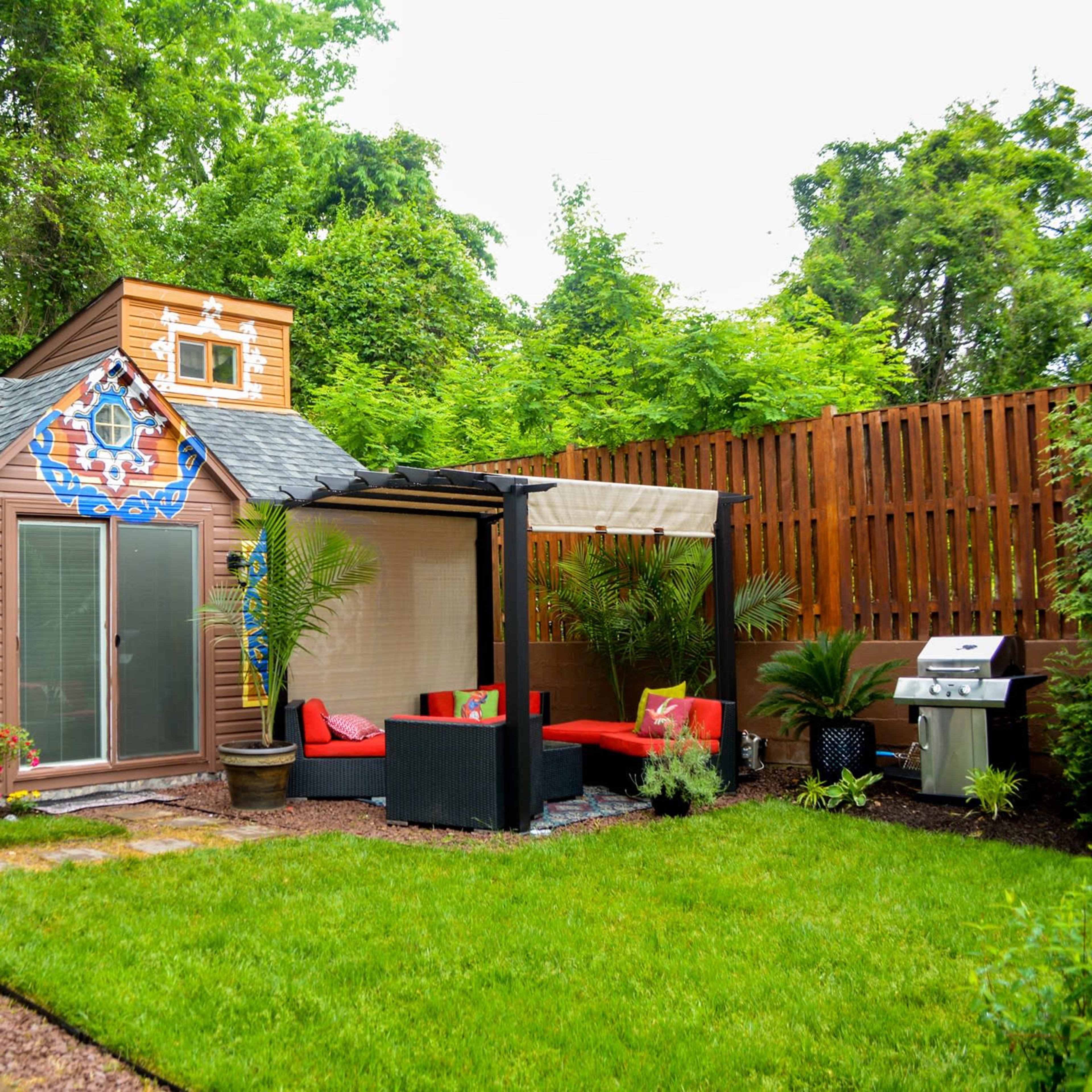 A small, colorful garden shed surrounded by greenery, with a patio area featuring red seating and a barbecue grill.