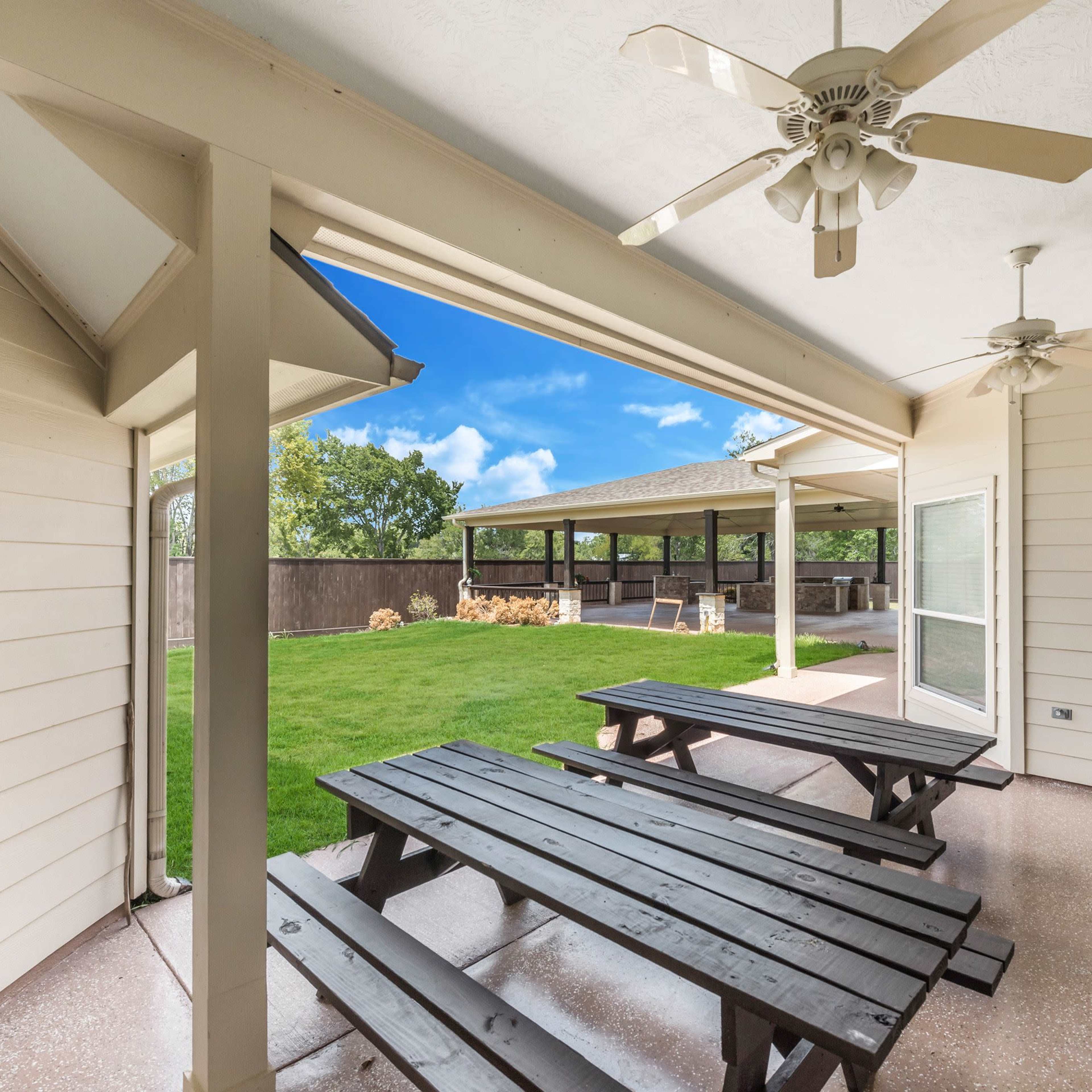 A covered outdoor patio area with multiple wooden picnic tables overlooking a lawn and a structure in the background.