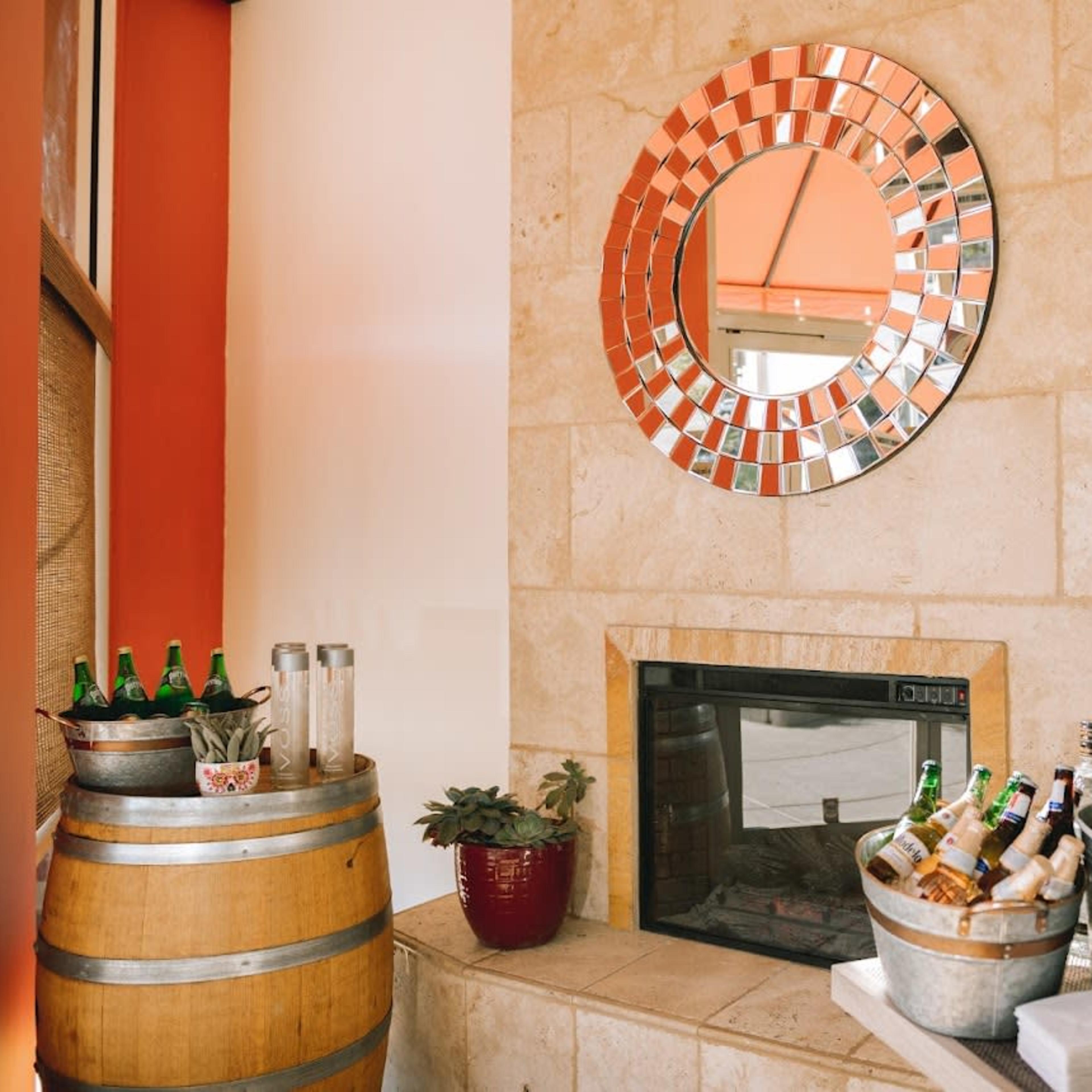 A cozy corner features a mirrored wall, a fireplace, a wine barrel, and a table with various bottles and drinks.