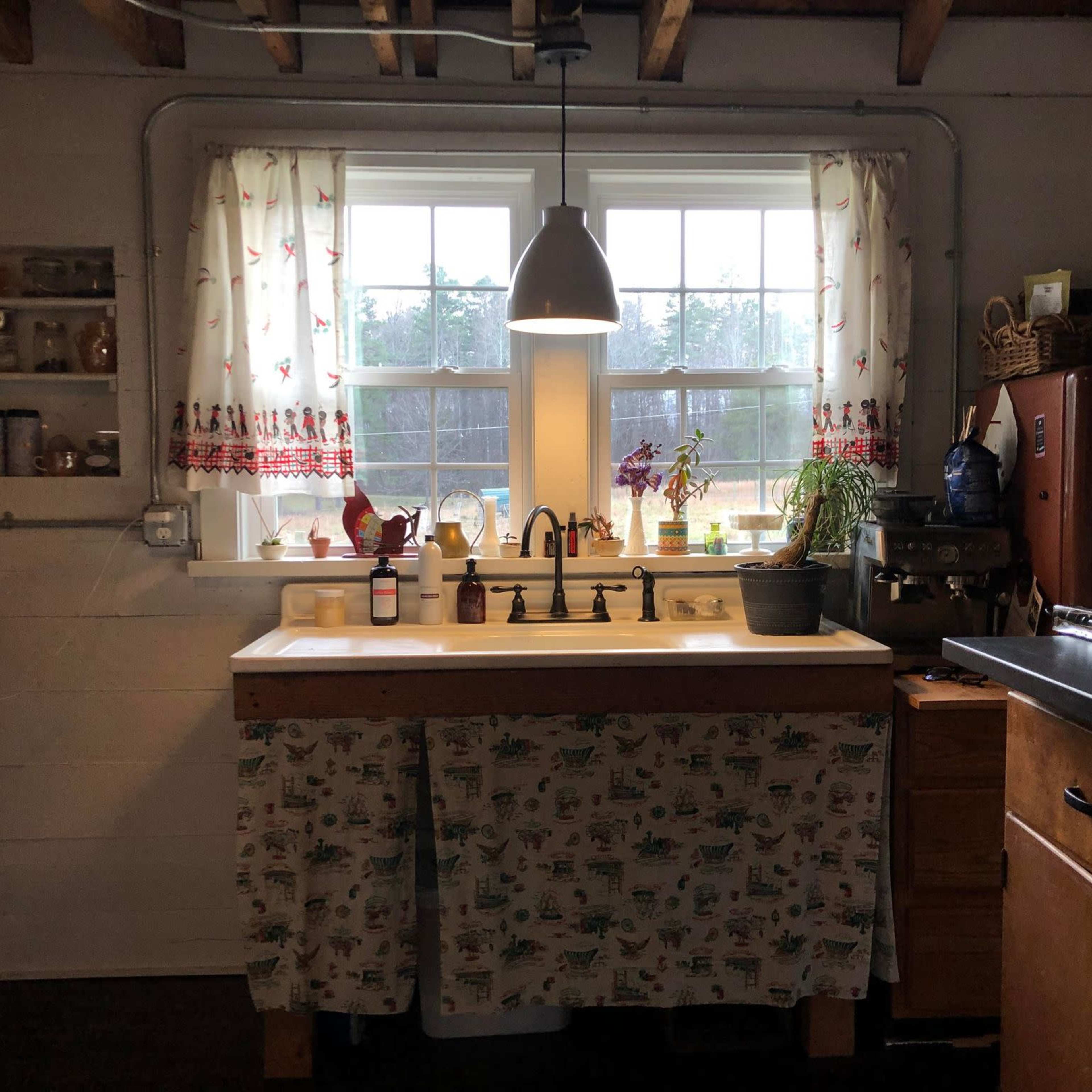 A kitchen with a white sink beneath two windows, a floral curtain, and a mix of modern and vintage appliances.