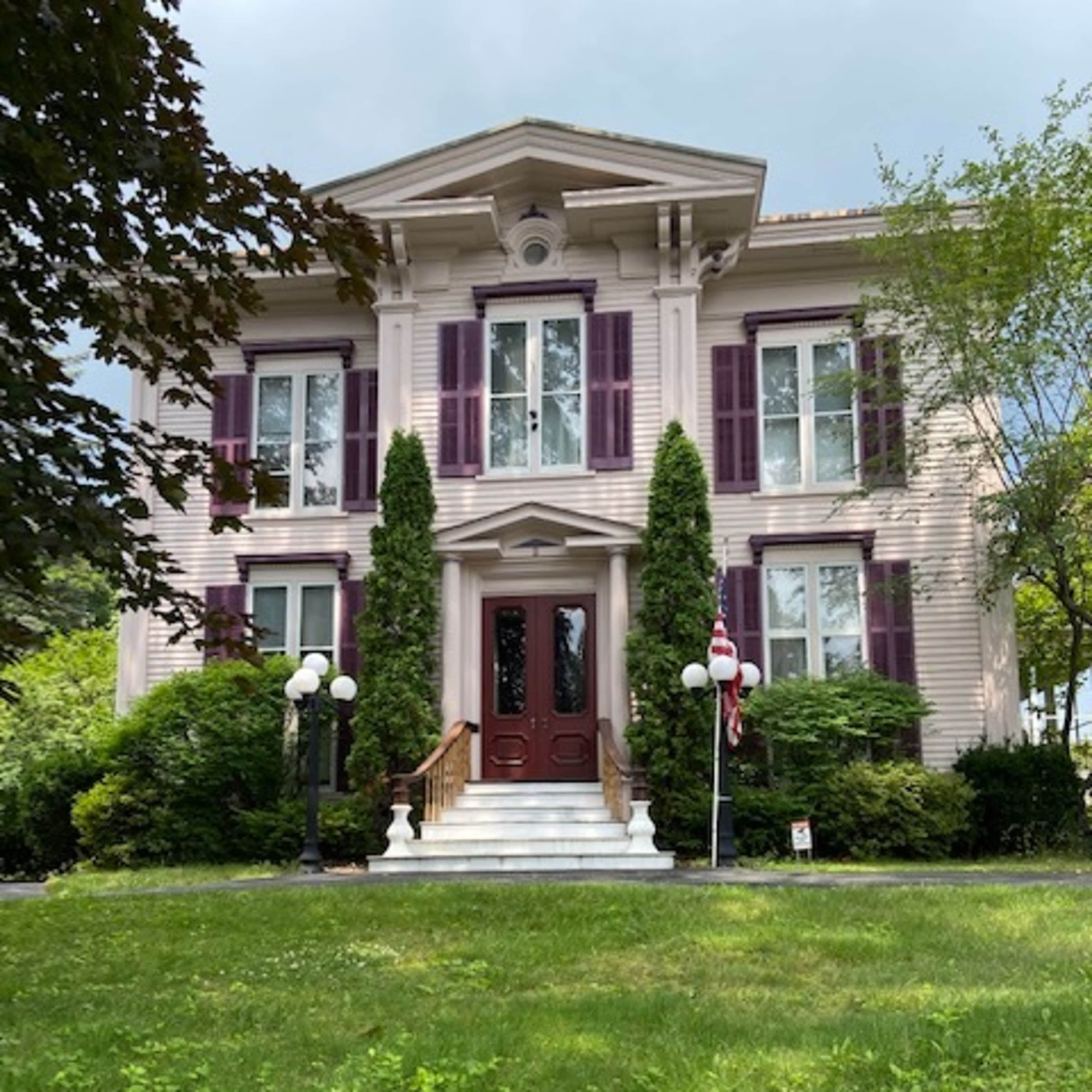 A large, two-story house with white siding, purple shutters, and a front porch flanked by tall, green shrubs.