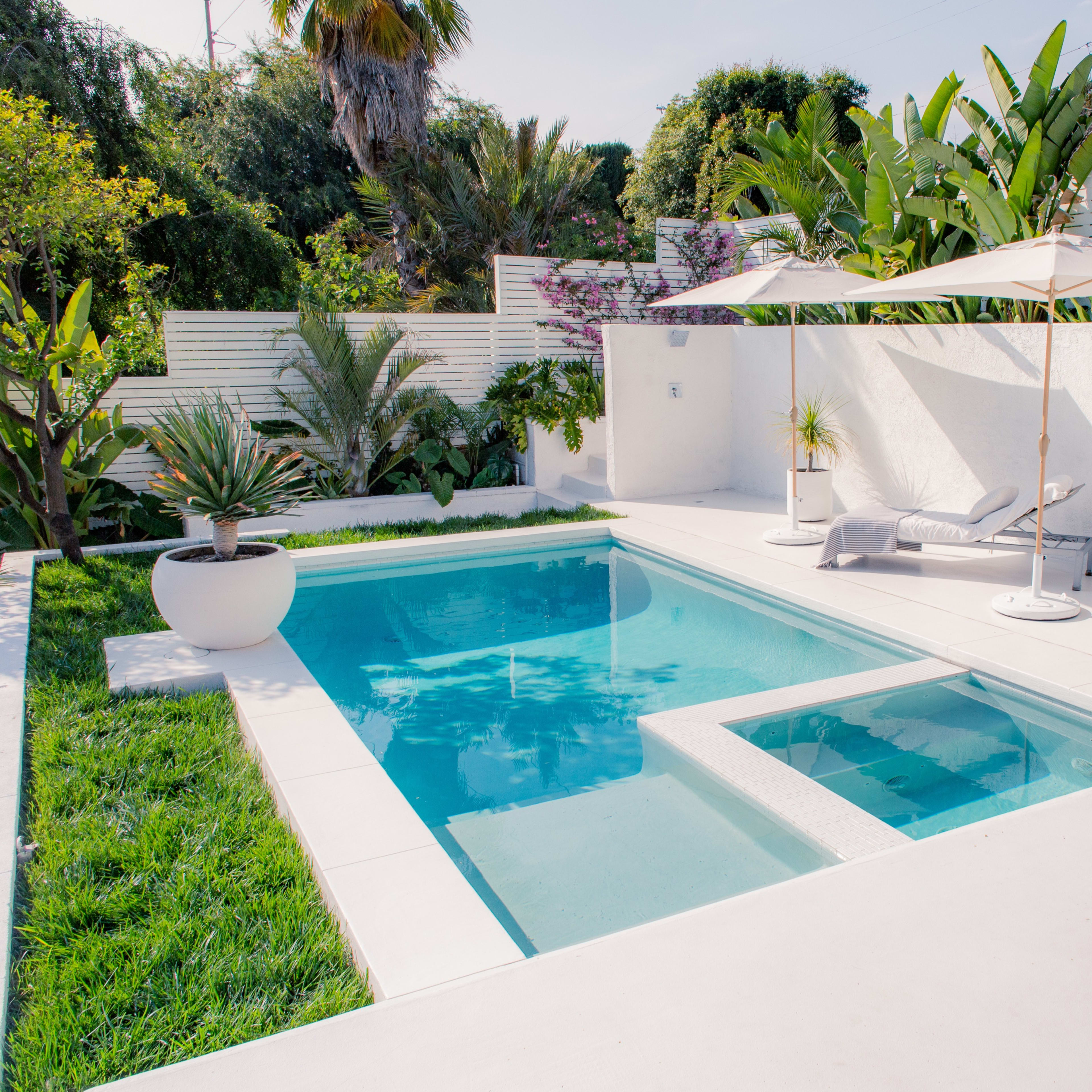 A rectangular swimming pool surrounded by white concrete surfaces, tropical plants, and a pair of lounge chairs with umbrellas.
