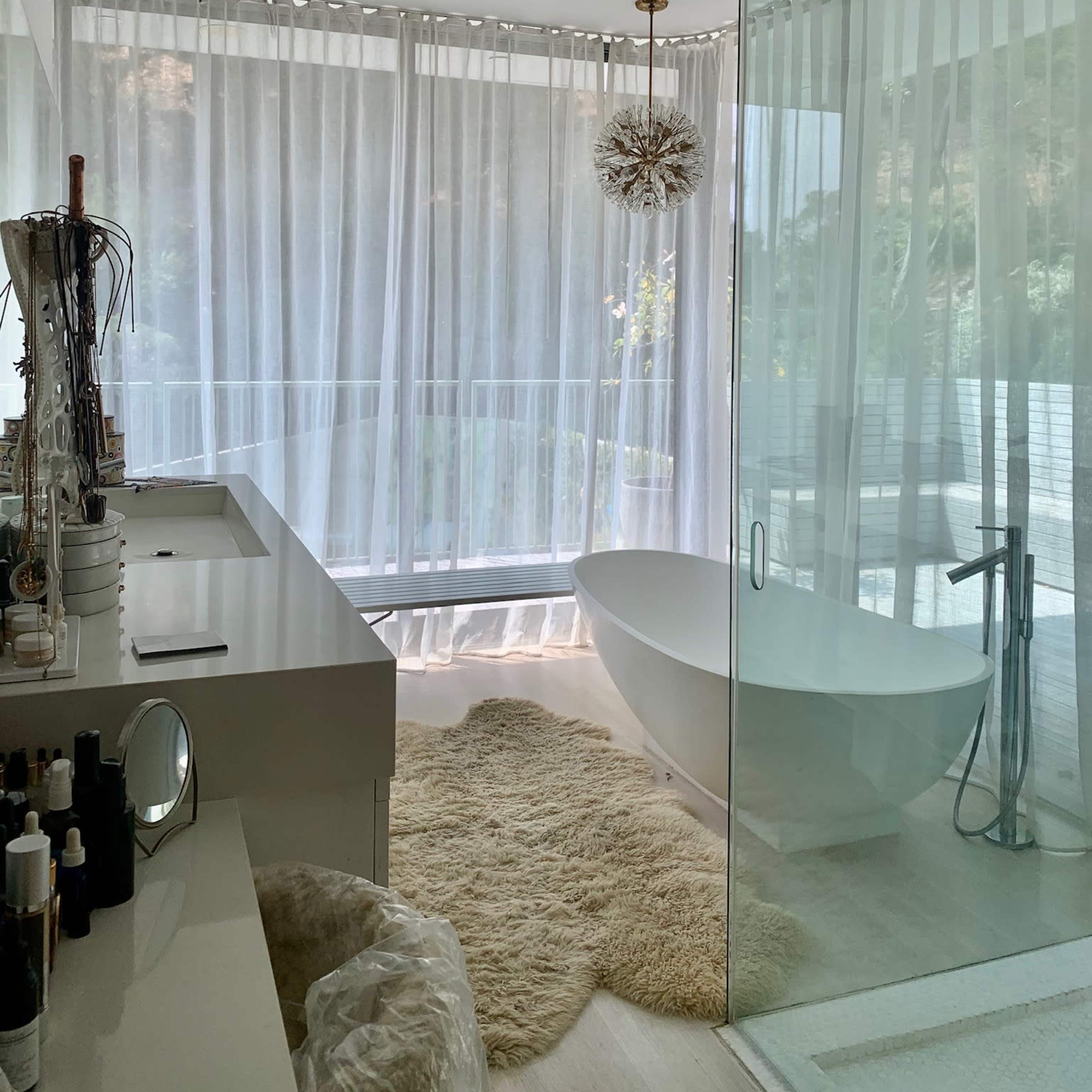 A modern bathroom features a freestanding tub, large windows covered with sheer curtains, and a fluffy area rug on the floor.