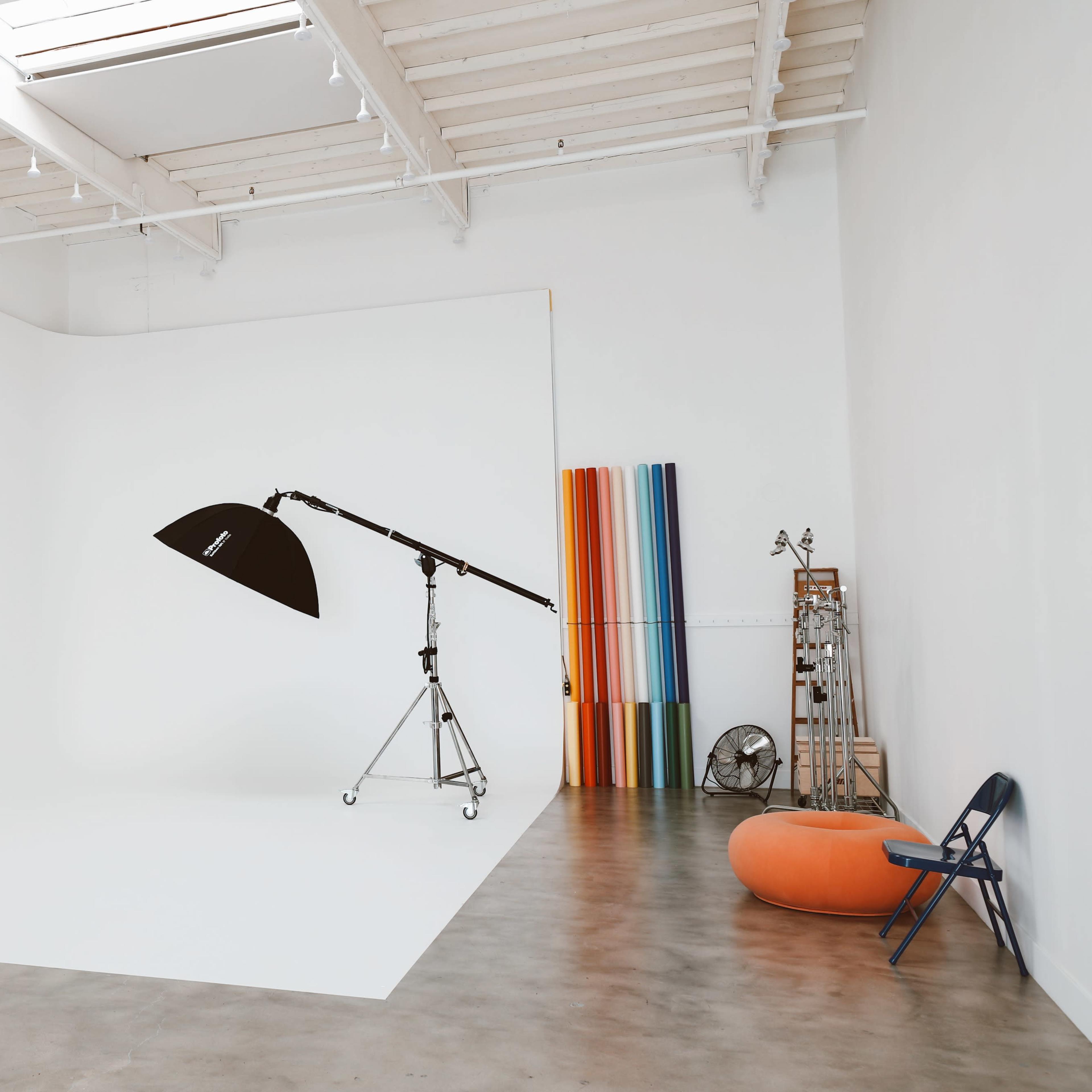 A spacious photography studio features a large white backdrop, a mounted light stand, and colorful backdrops neatly arranged along the wall.