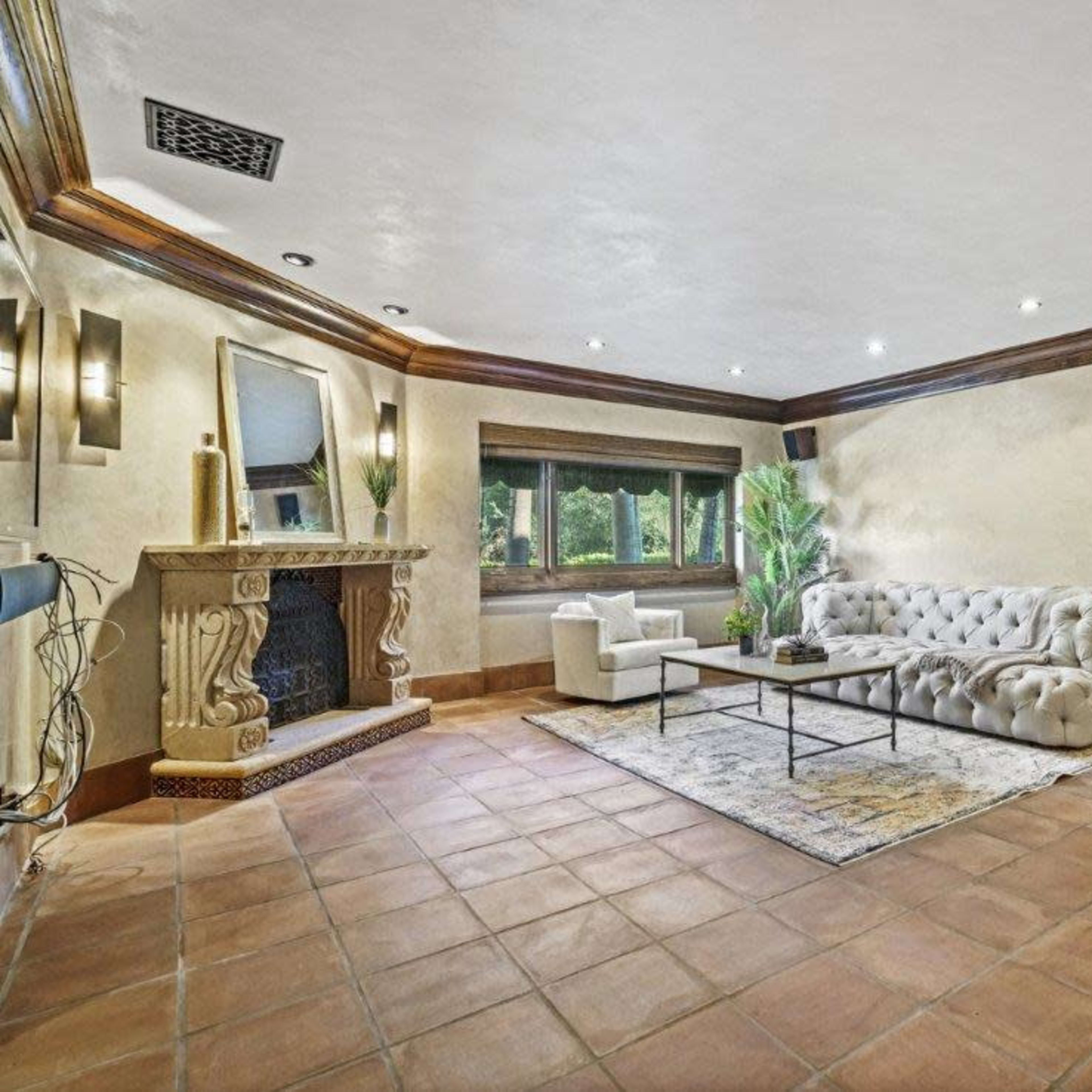 The room features a light-colored wall with a decorative fireplace, a large tufted sofa, a coffee table, and a large window allowing natural light to enter.