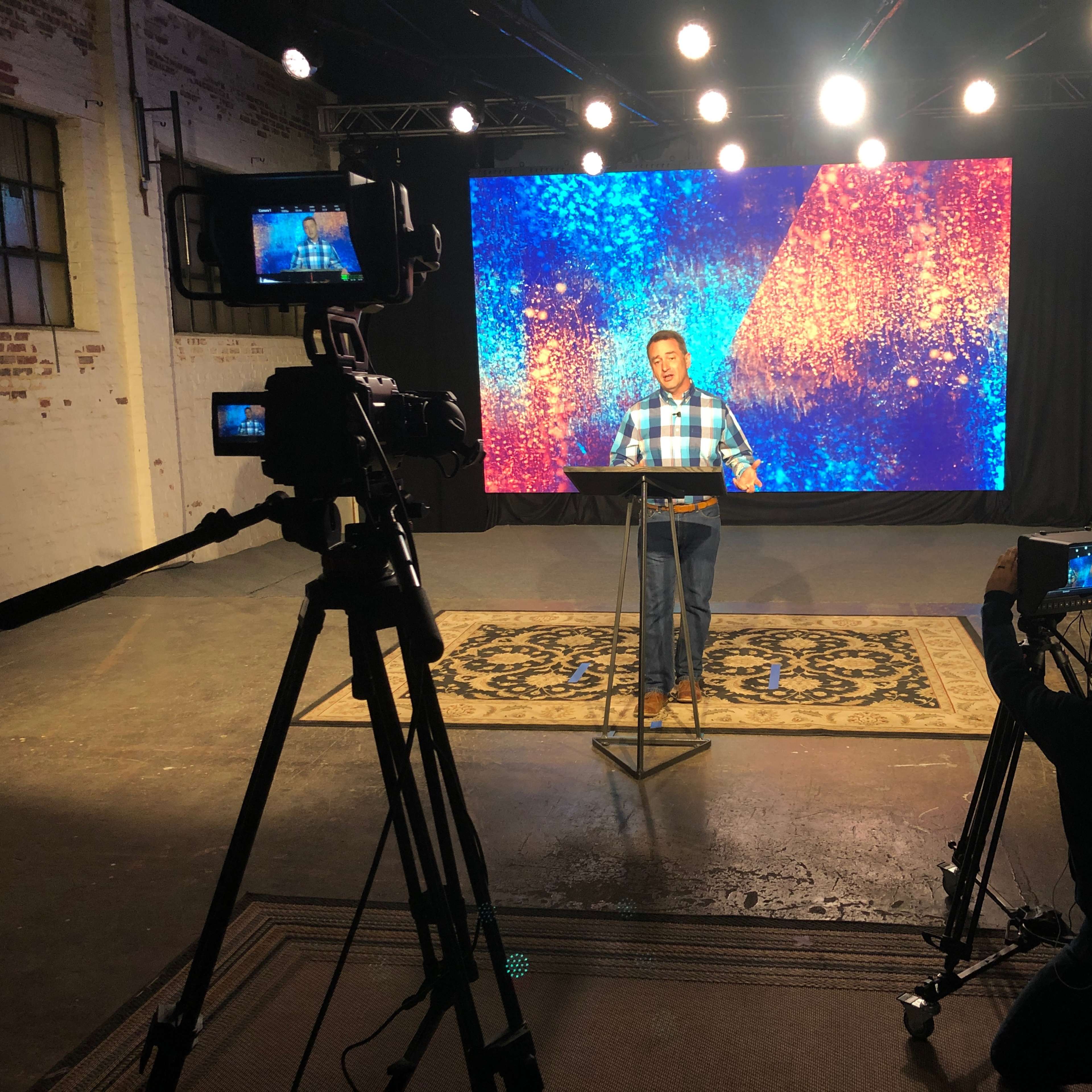 A speaker stands at a podium in front of a colorful backdrop with multiple cameras set up to capture the presentation.