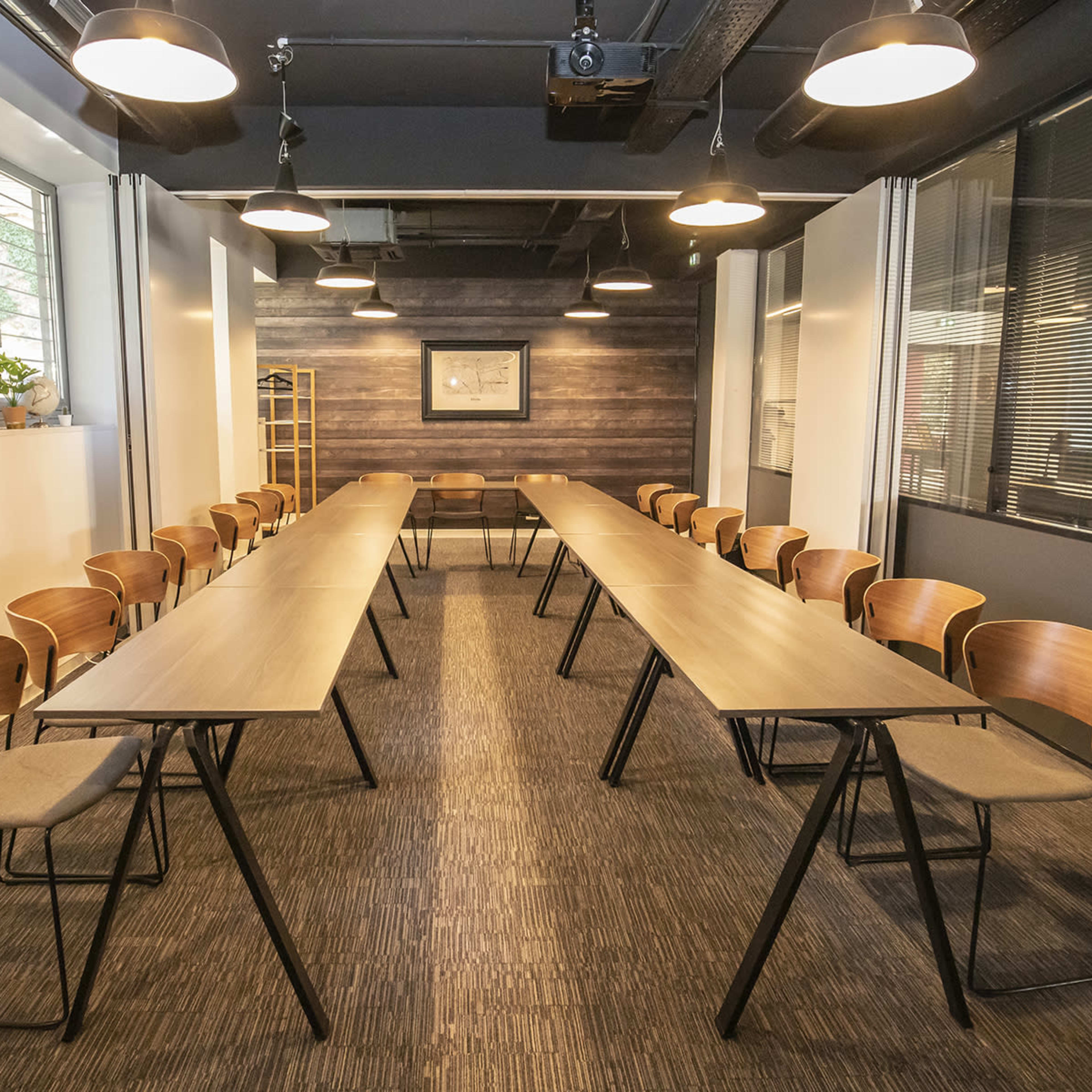 A conference room is set up with two rows of tables and chairs facing each other, surrounded by large windows and diverse wall textures.