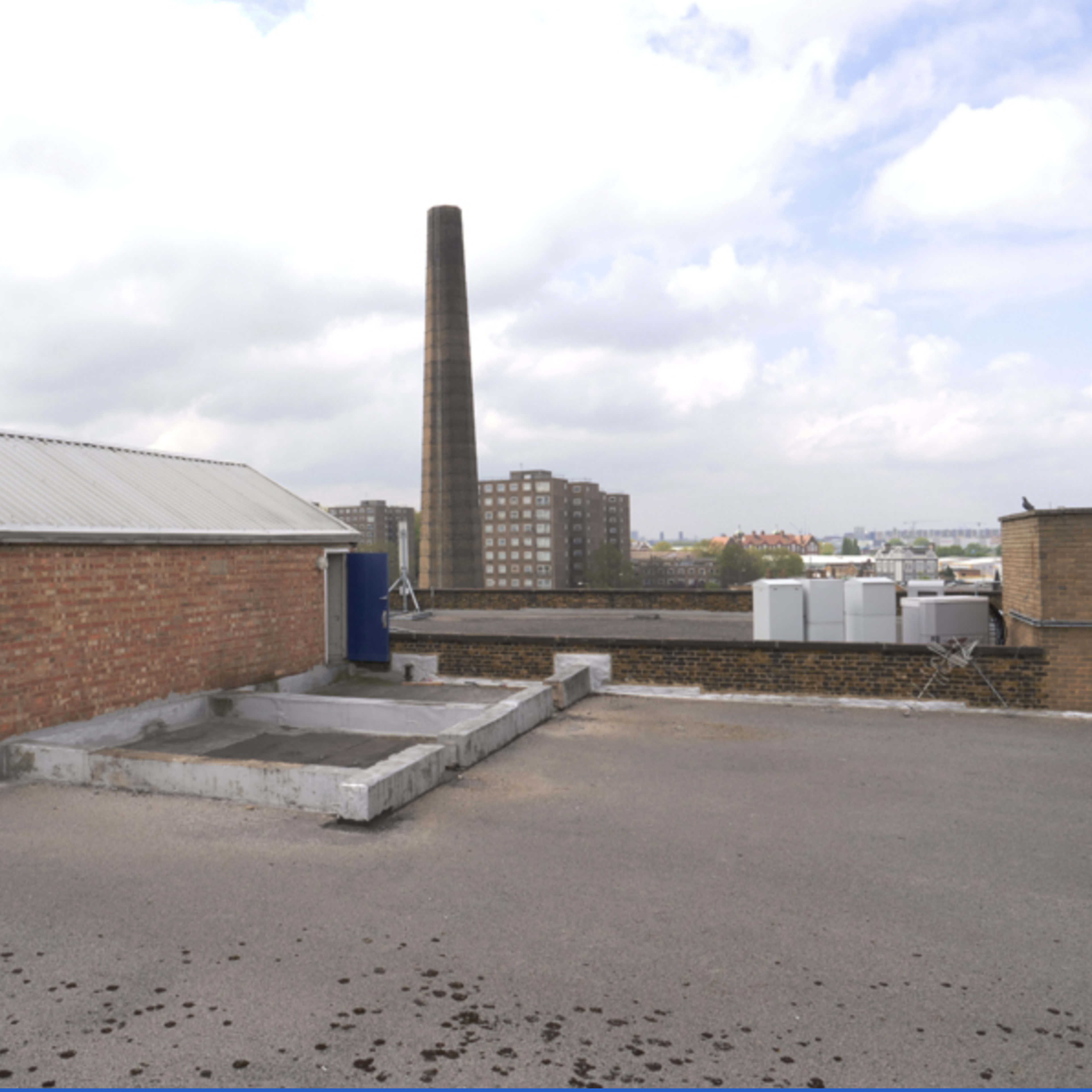 A rooftop with a brick building and a tall smokestack in the background, alongside several low-rise buildings and a cloudy sky.