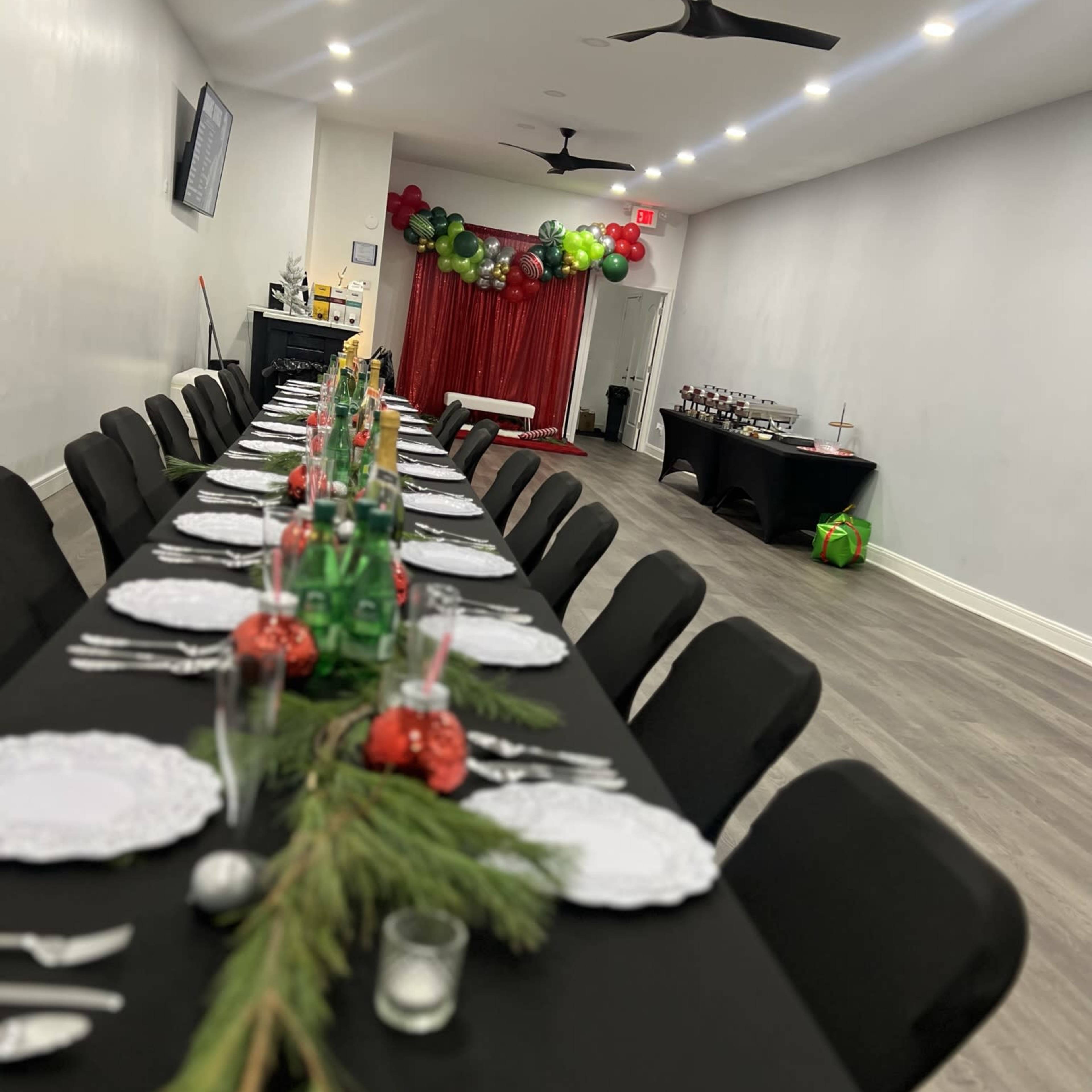 A long table is set for a festive event with green and red decorations, featuring plates, glasses, and a centerpiece of greenery.