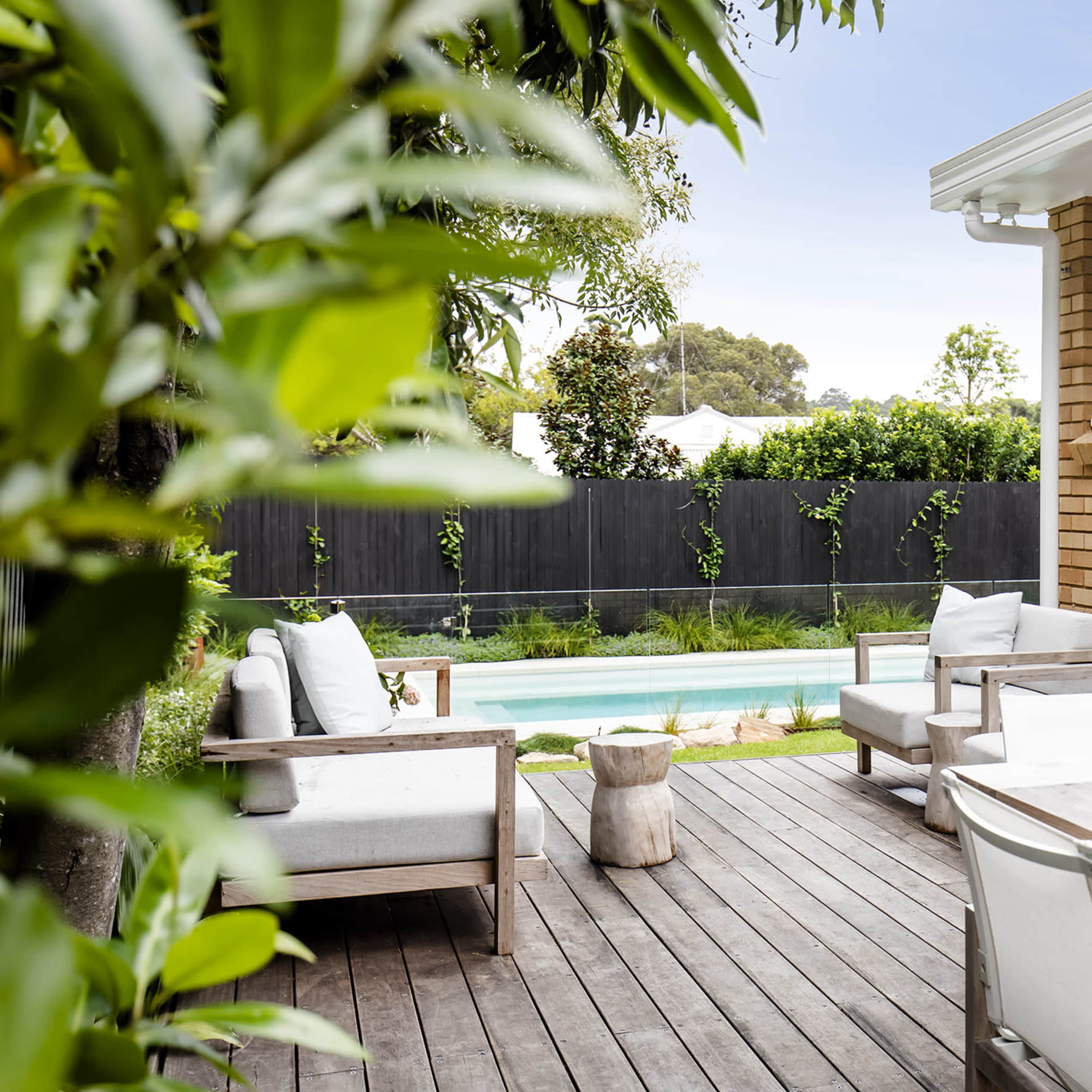 A wooden deck features a table and chairs alongside two lounge chairs, with a green lawn and a pool visible in the background.