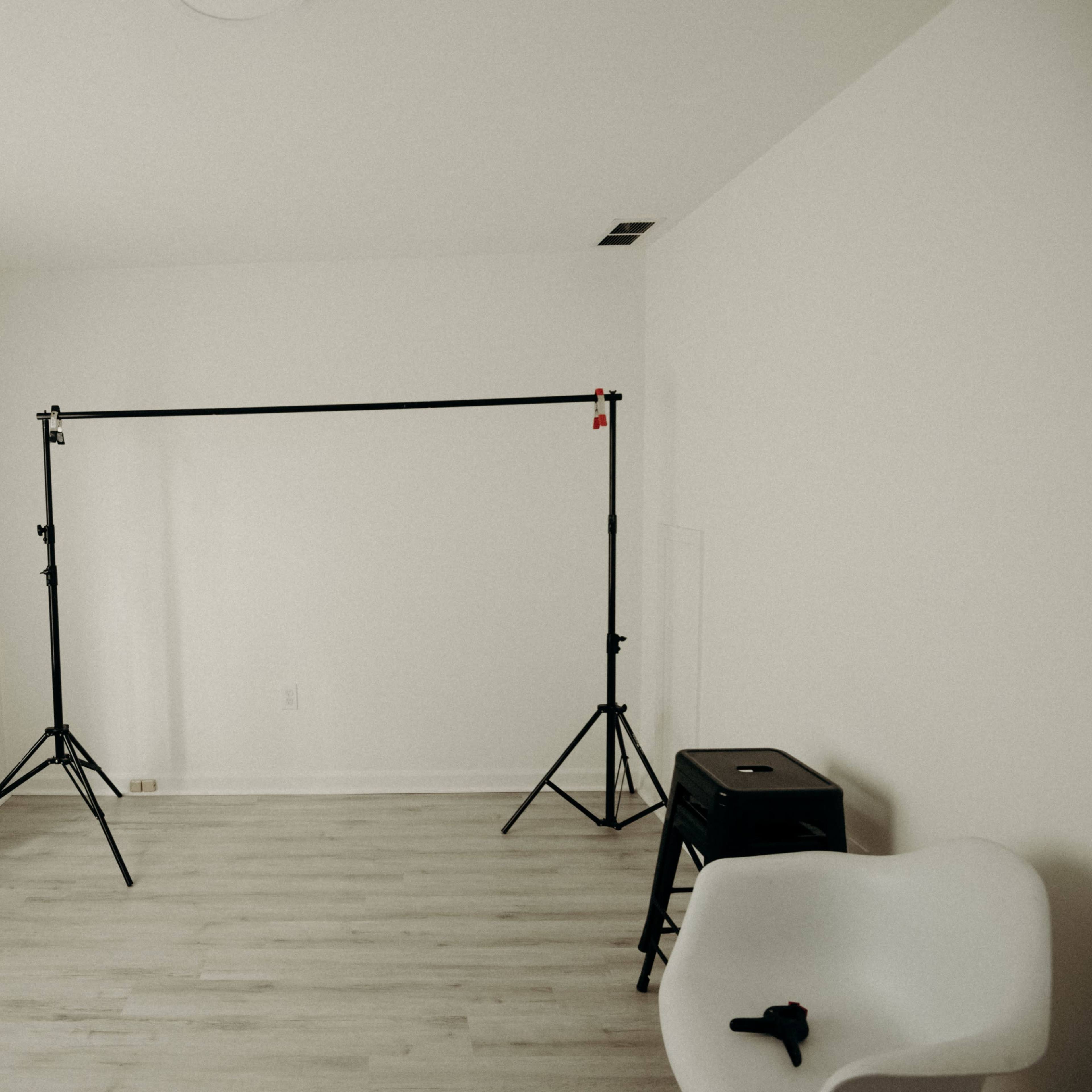 A minimalistic room with light wooden flooring, a white wall, natural light coming through sheer curtains, and a black backdrop stand with tripods.