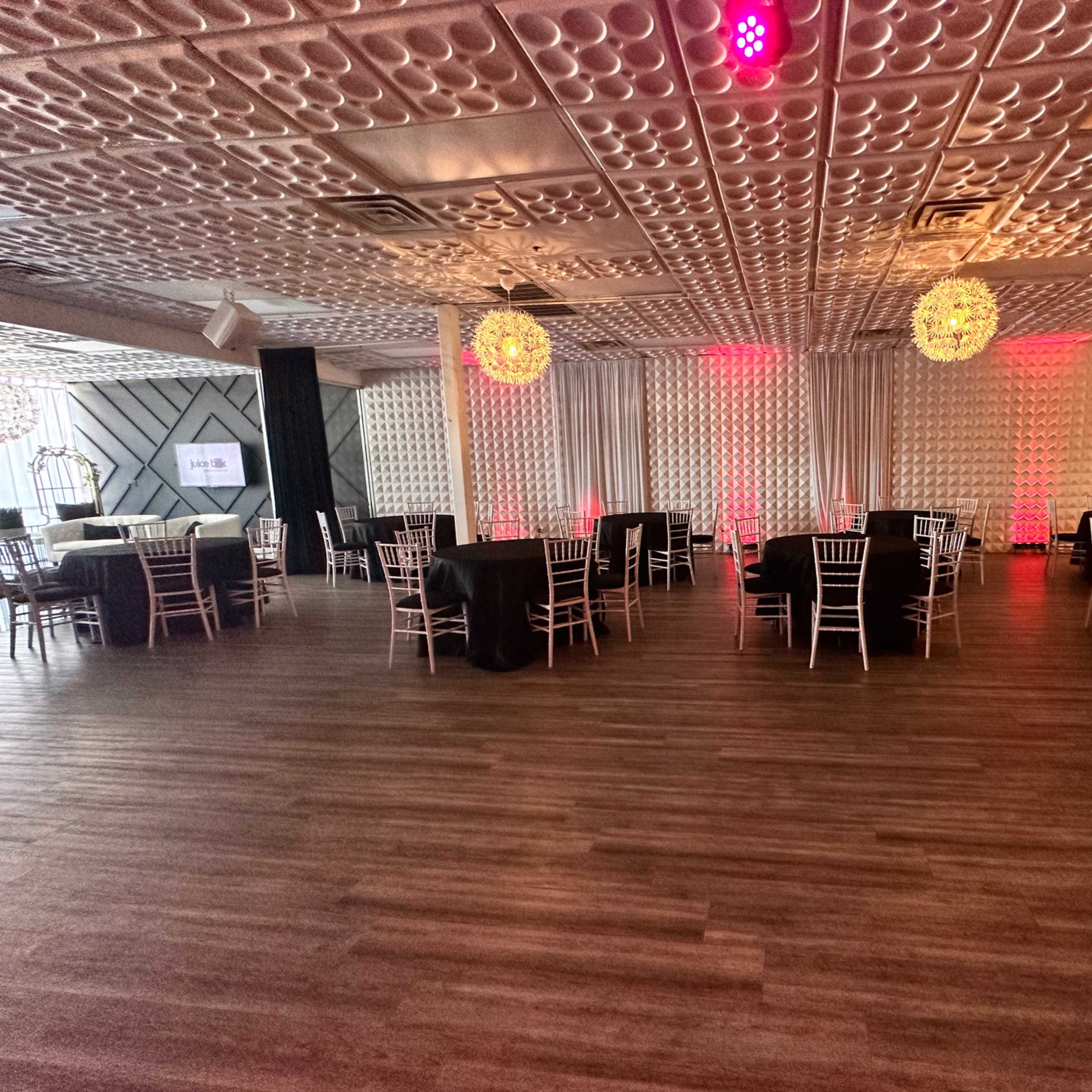 A spacious event venue set up with round tables covered in black tablecloths and elegant chairs, featuring decorative lighting and a bar area in the background.