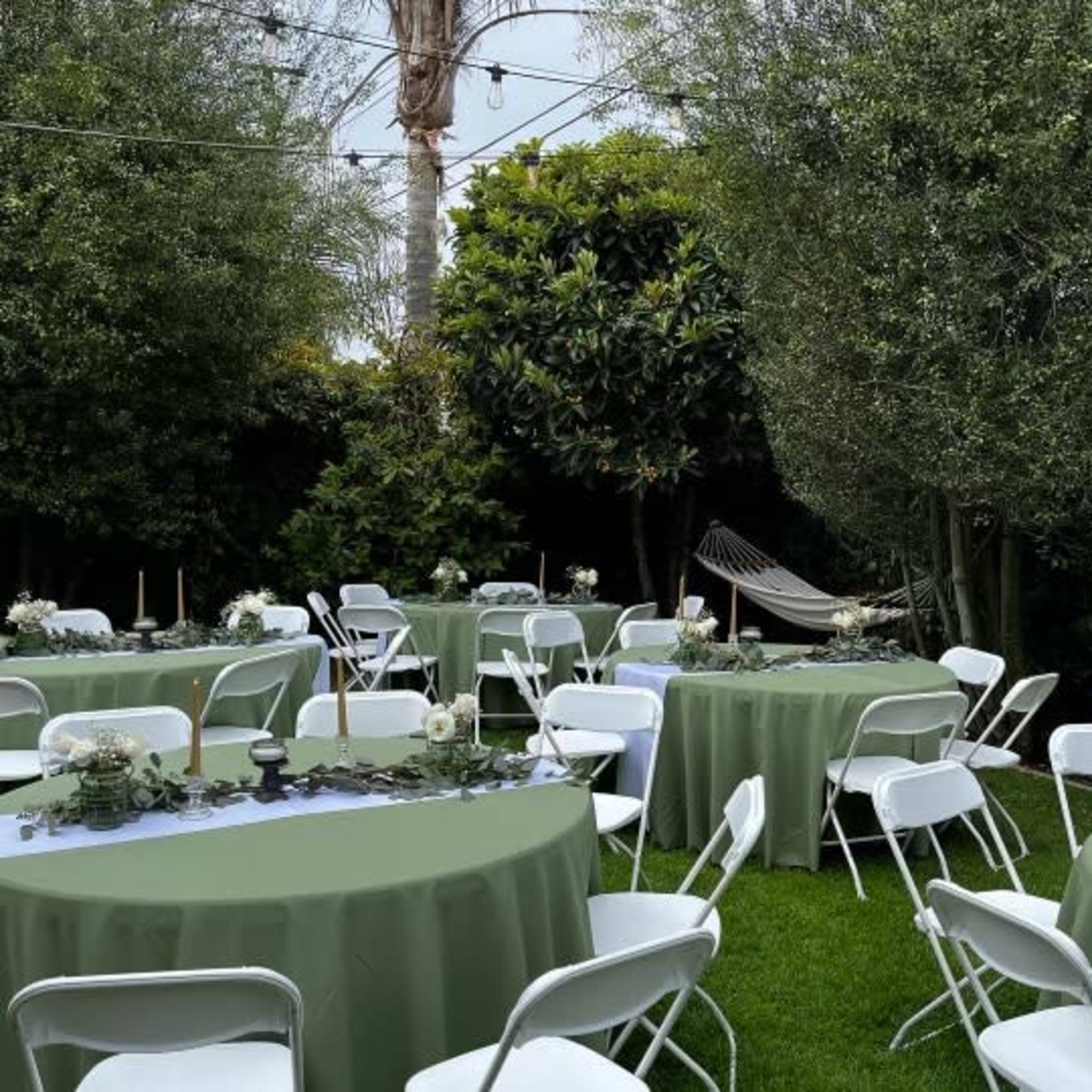 A garden setting with round tables covered in green tablecloths, surrounded by trees and a hammock in the background.