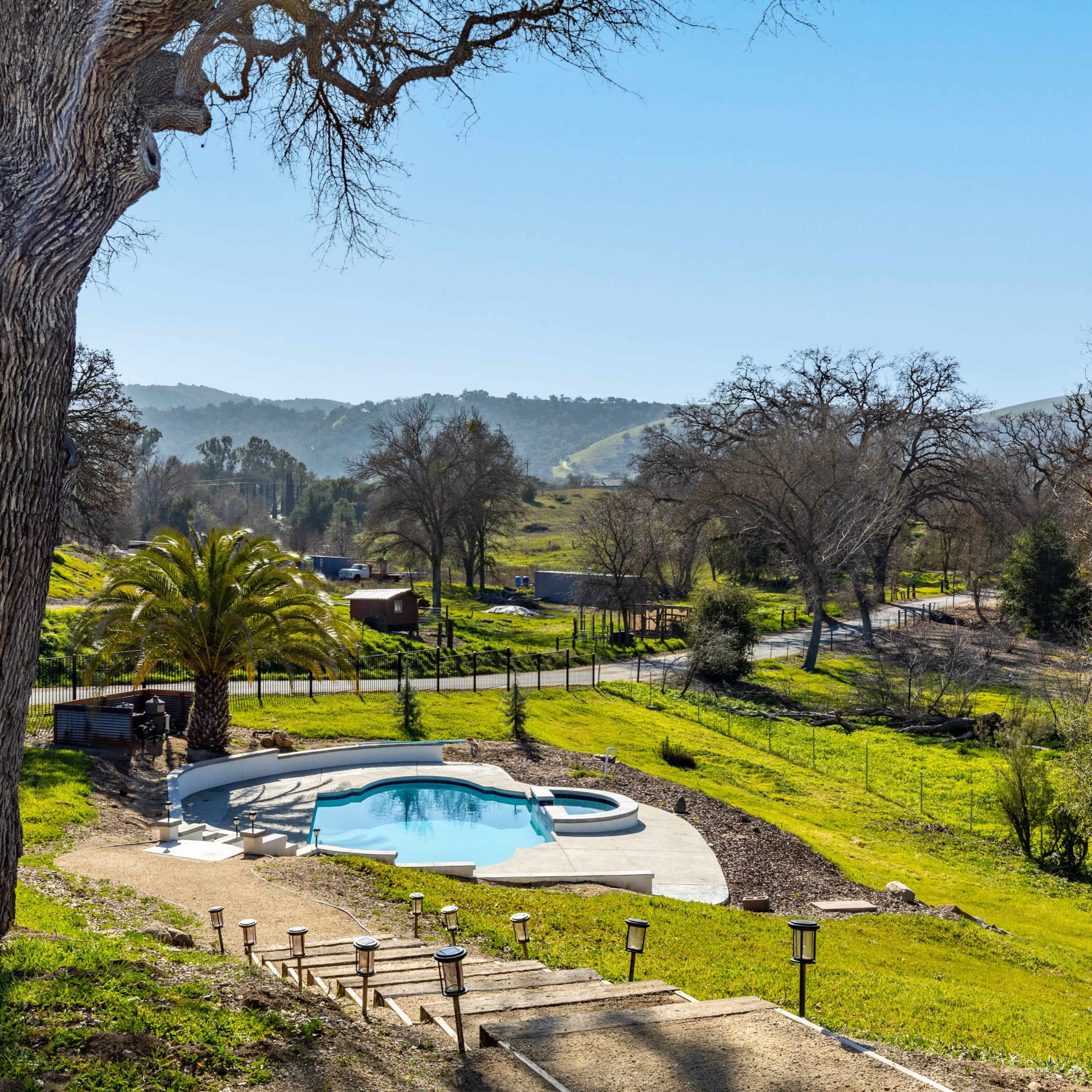 A swimming pool is situated on a grassy hillside, surrounded by trees and hills in the background.
