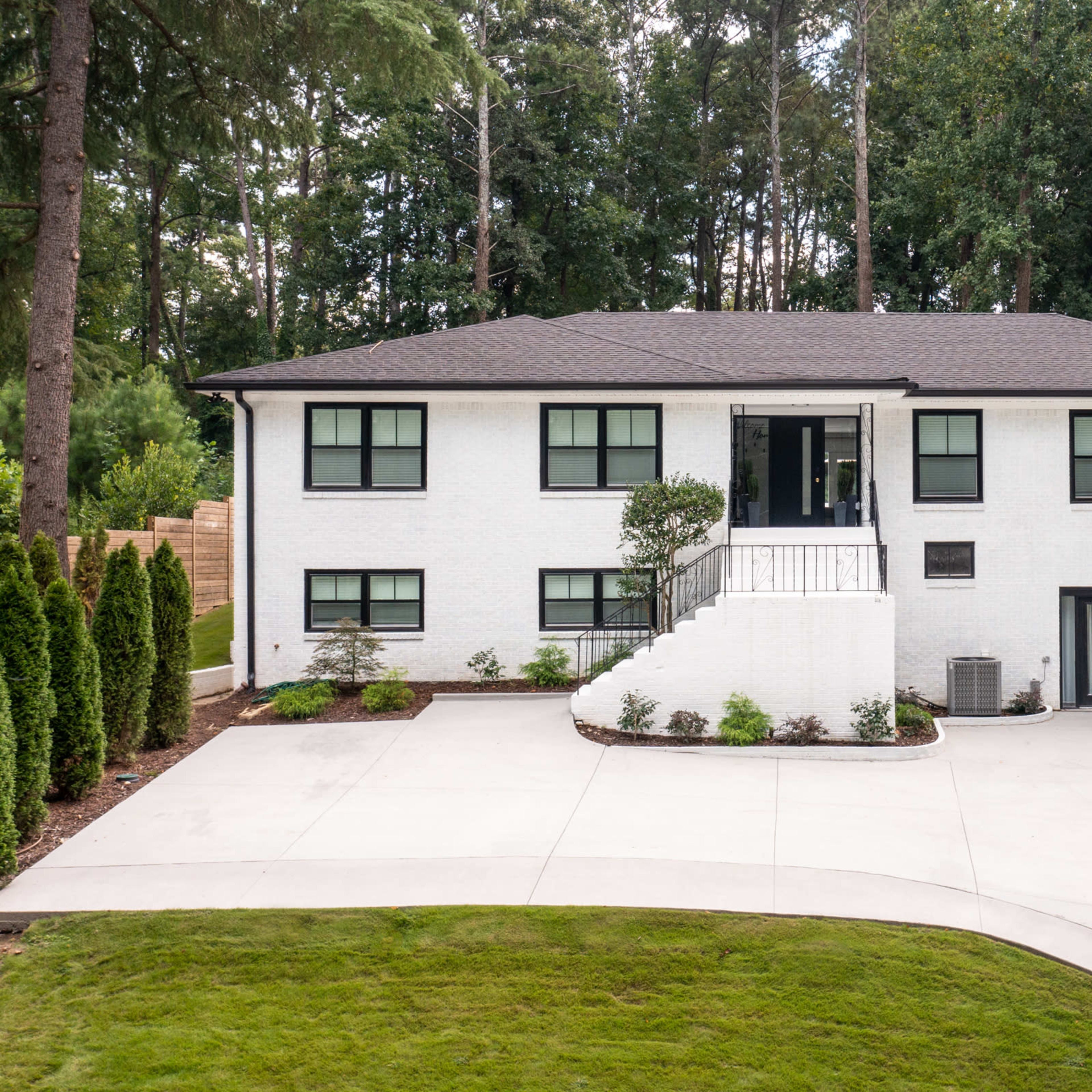 A modern two-story white house is surrounded by greenery with a large concrete driveway leading up to it.