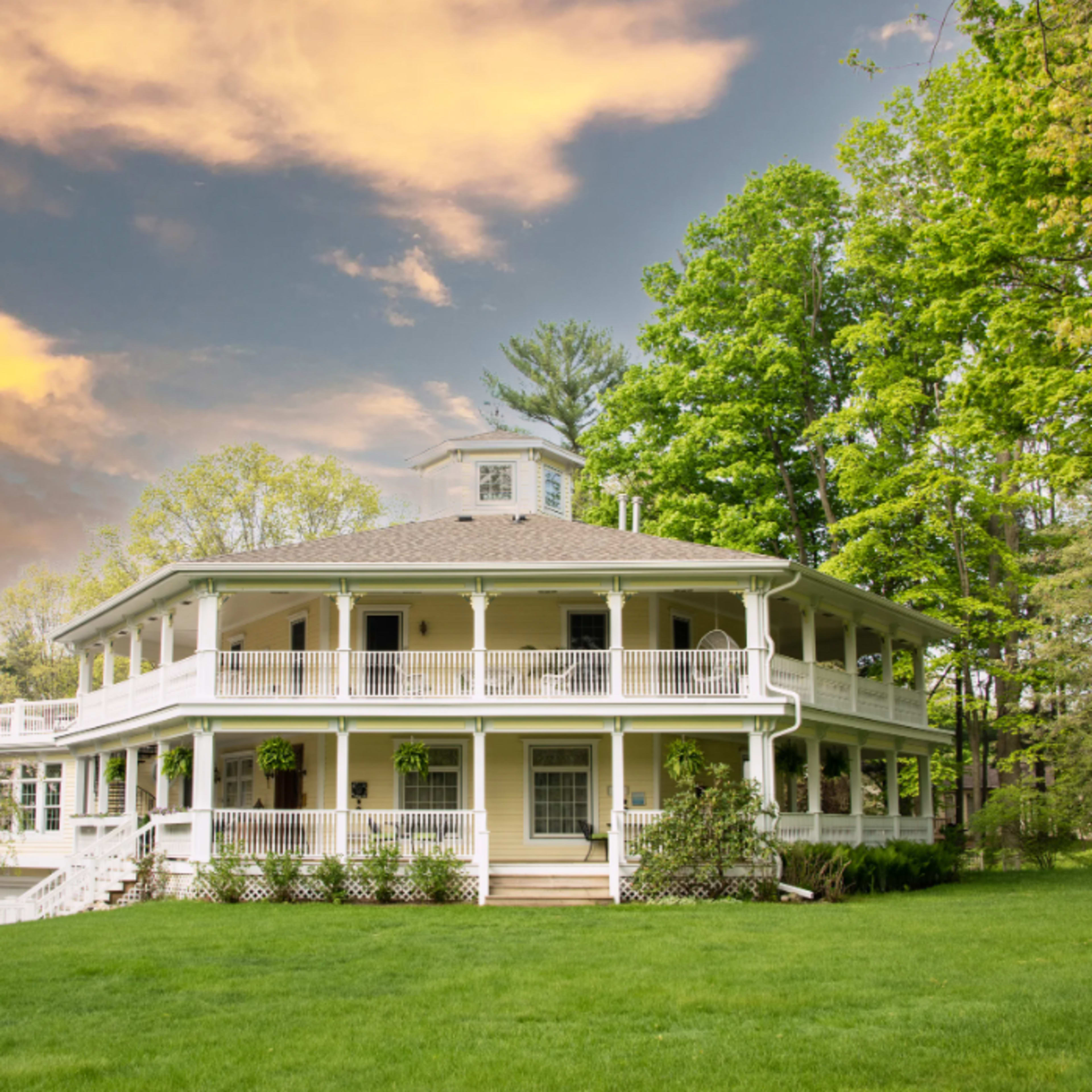 A large, two-story white house with a circular design, multiple balconies, and surrounded by greenery under a cloudy sky.