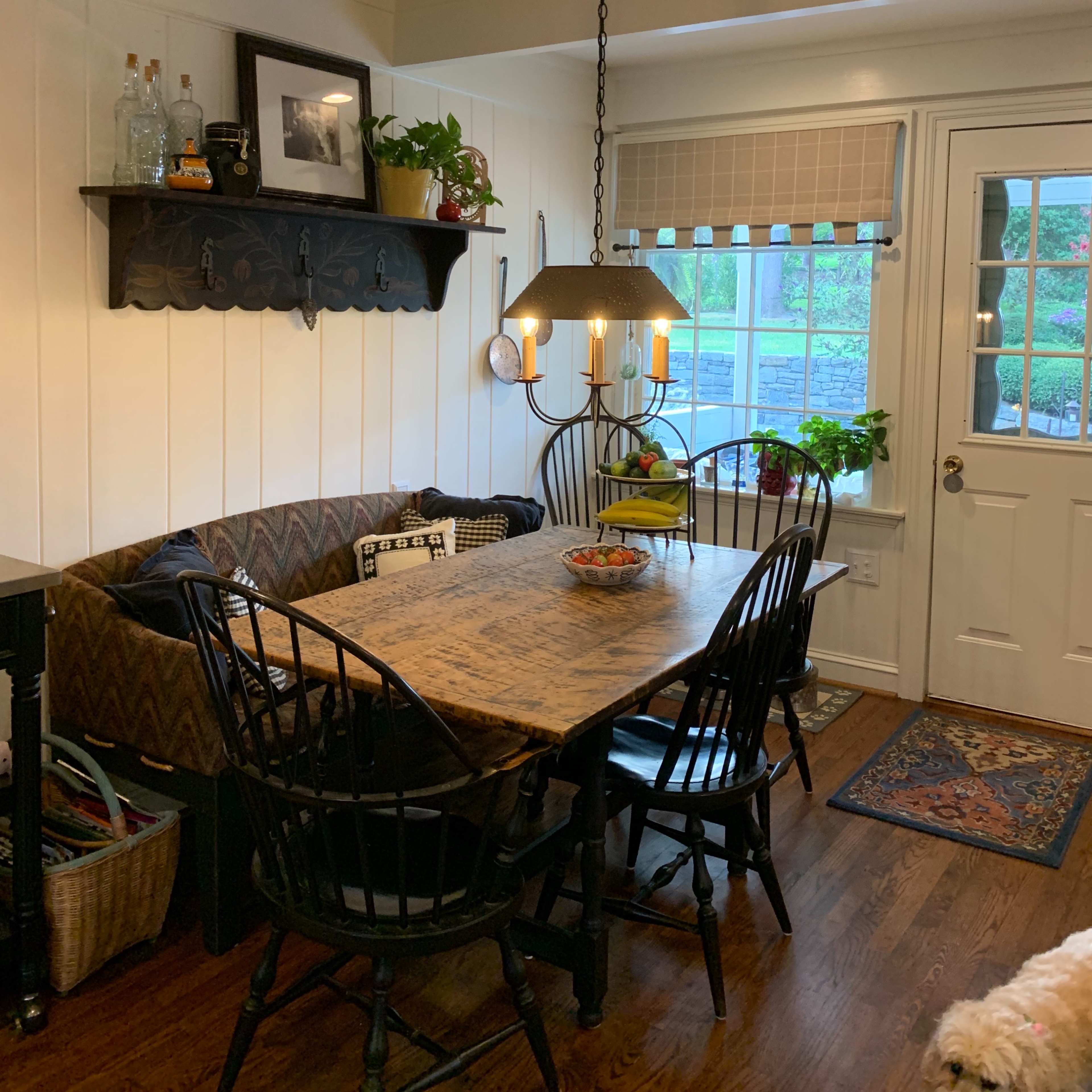 A cozy kitchen nook features a wooden table surrounded by black chairs, a built-in bench with patterned cushions, and a large window beside a door.