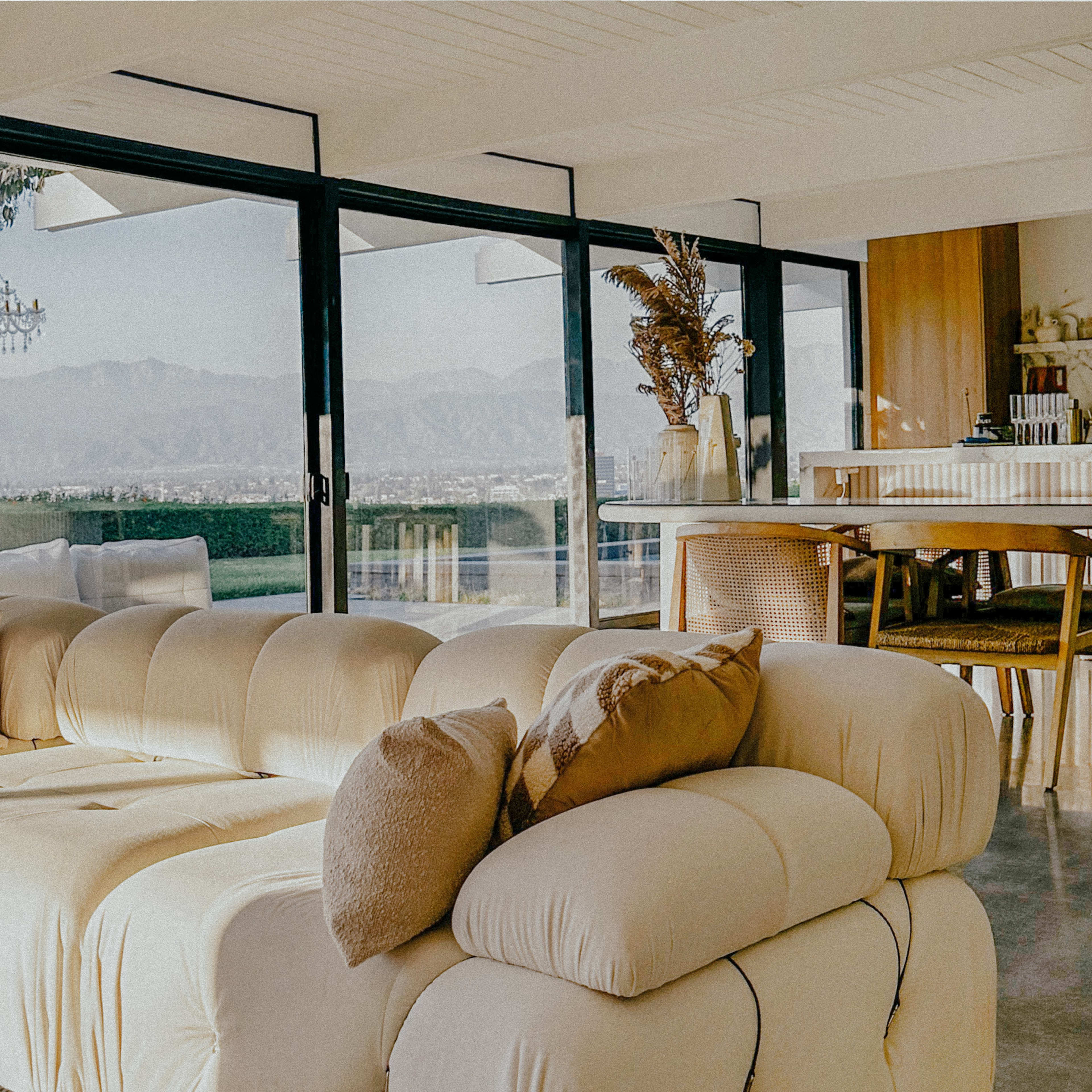 A spacious living area features a white, plush sofa with decorative pillows, large glass windows showcasing a mountain view, and a modern dining space nearby.