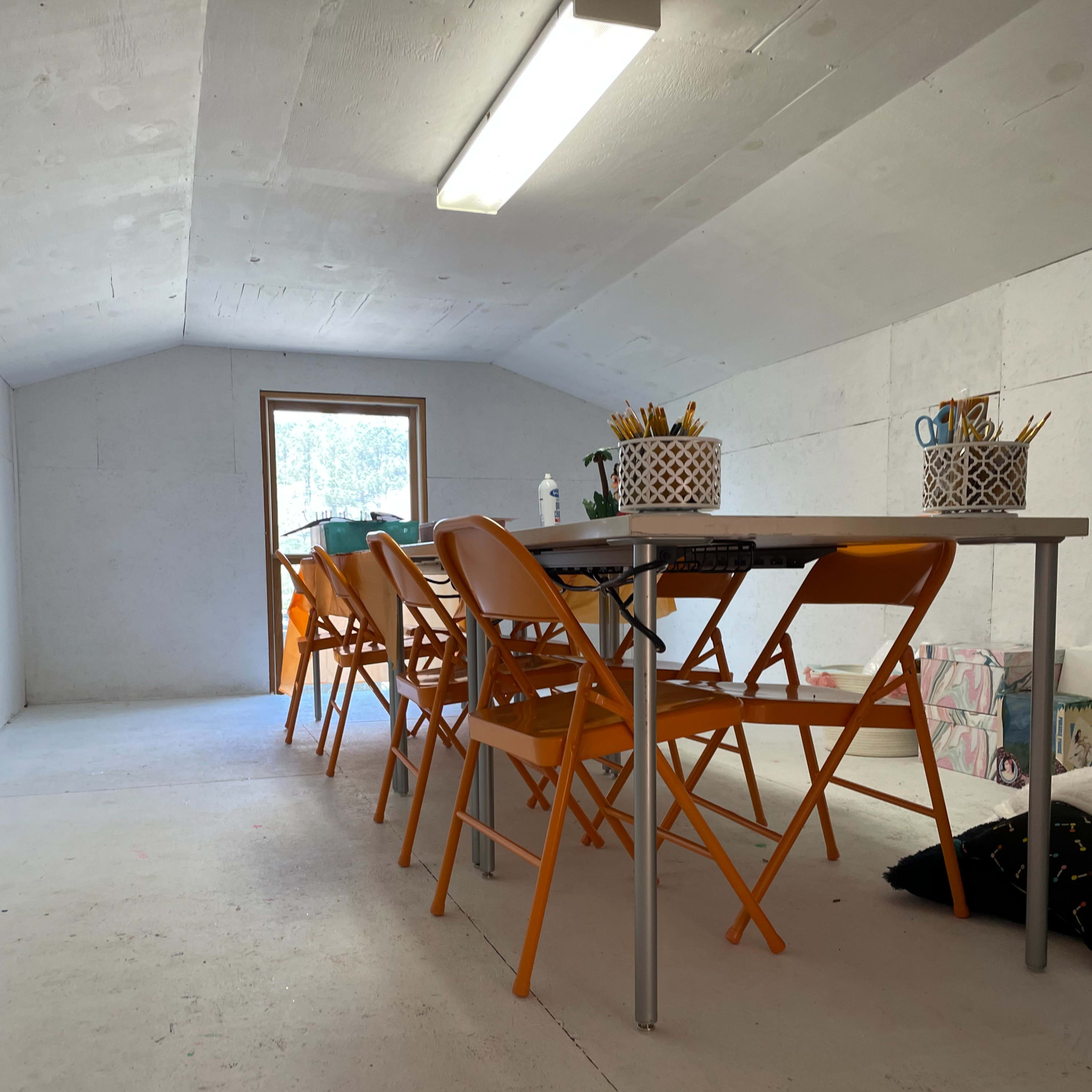 A spacious, light-filled attic room features a long table surrounded by orange folding chairs.