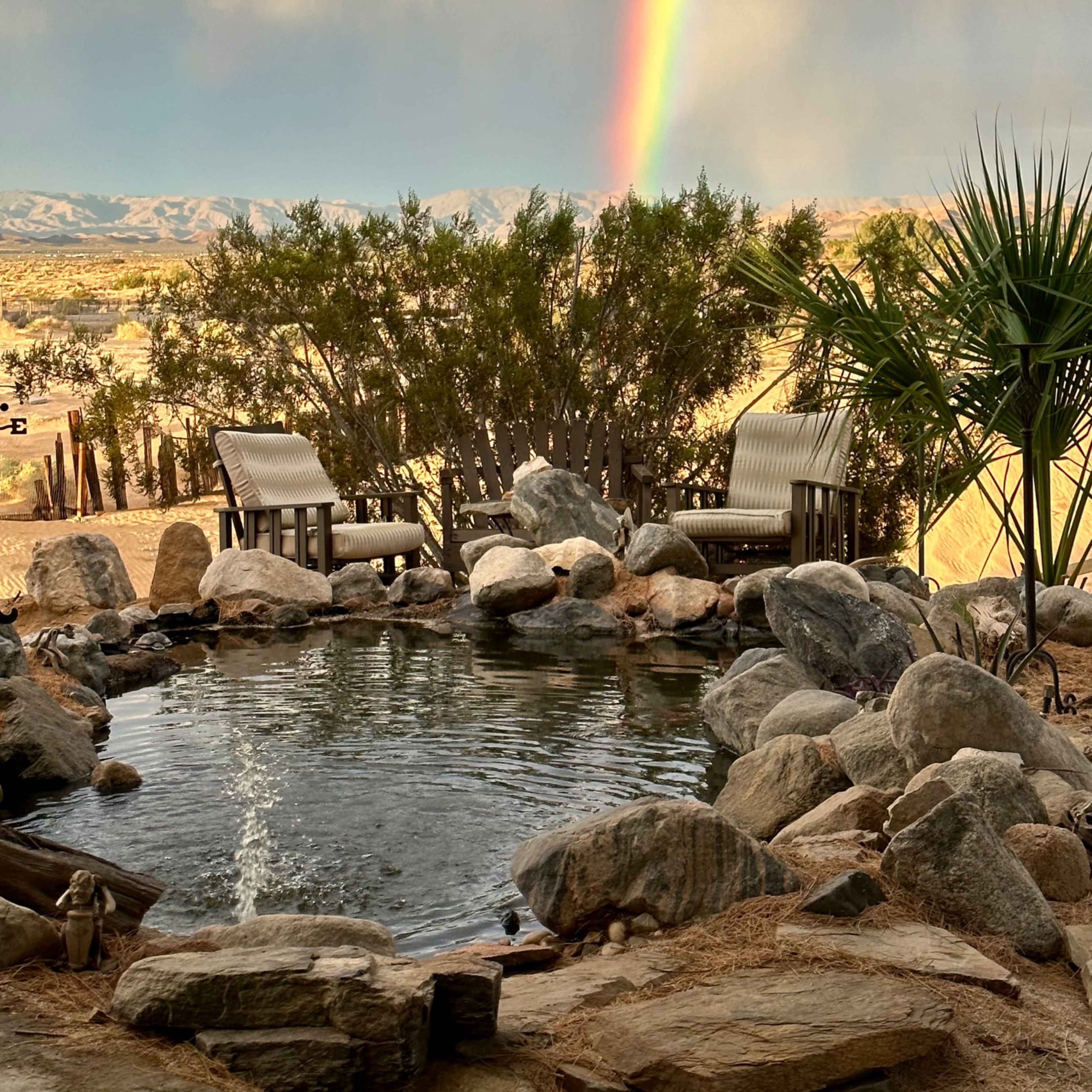 A small pond surrounded by rocks and two chairs sits in a desert landscape, with a rainbow arching in the background.