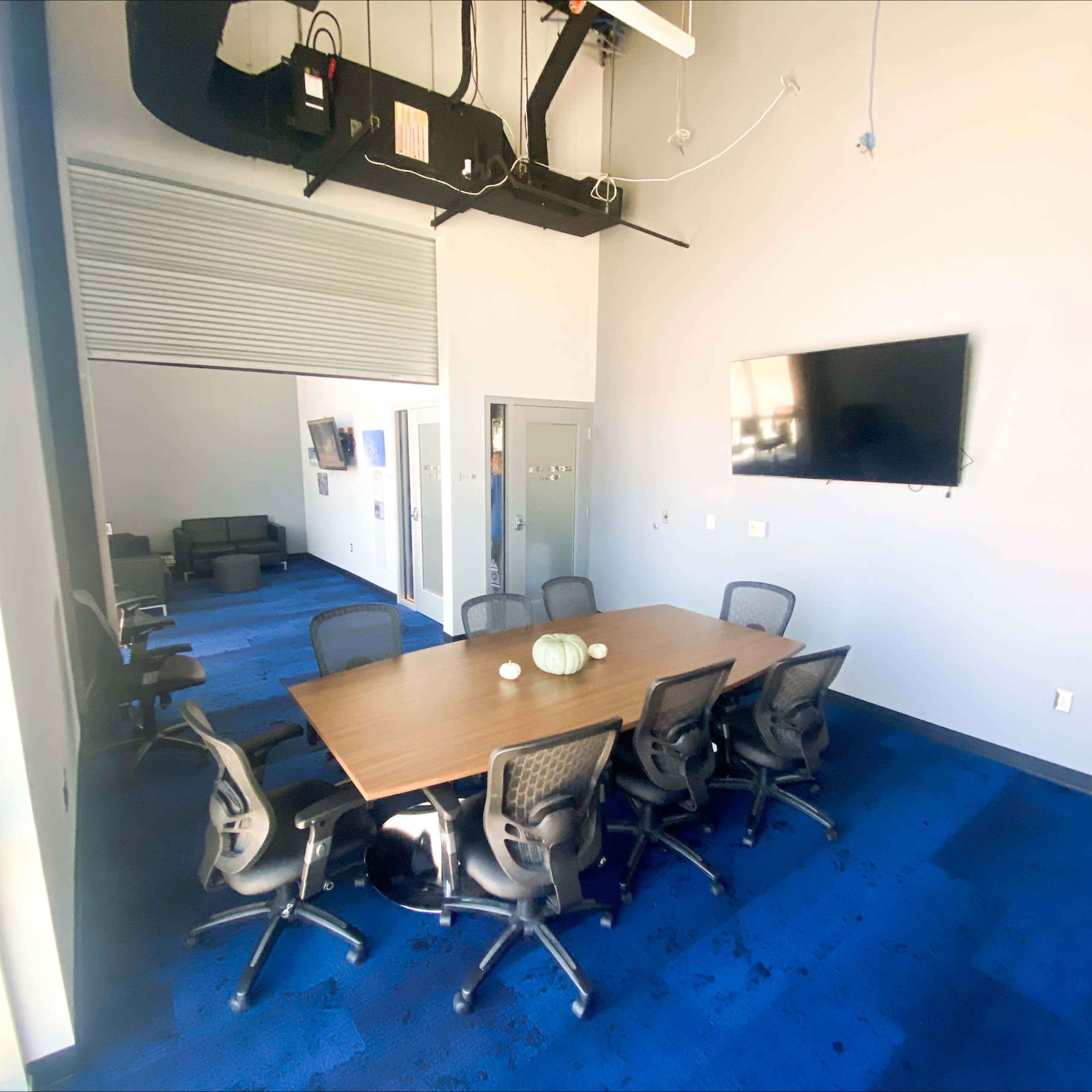 A modern conference room features a large wooden table surrounded by ergonomic chairs, with a wall-mounted screen and a couch visible in the background.