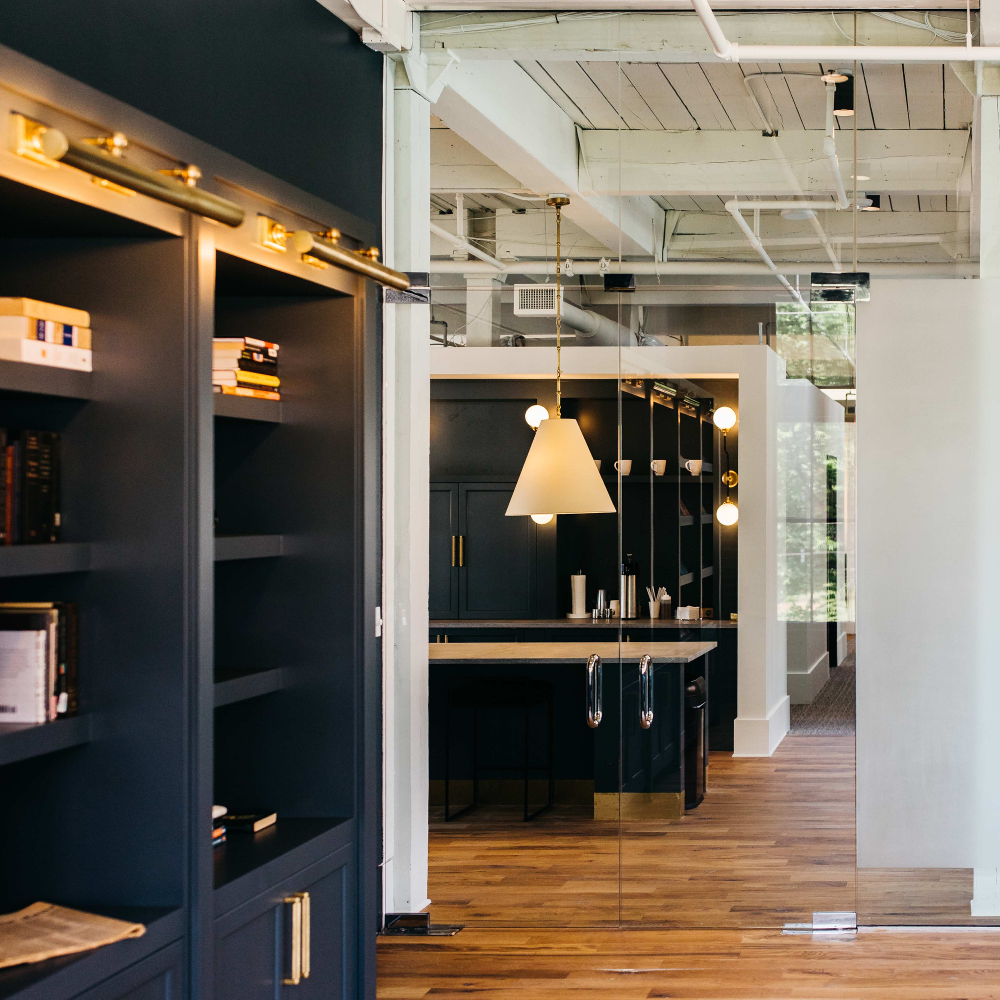 A modern office space featuring dark cabinetry, wooden flooring, and a mirrored wall leading to a kitchen area with pendant lighting.