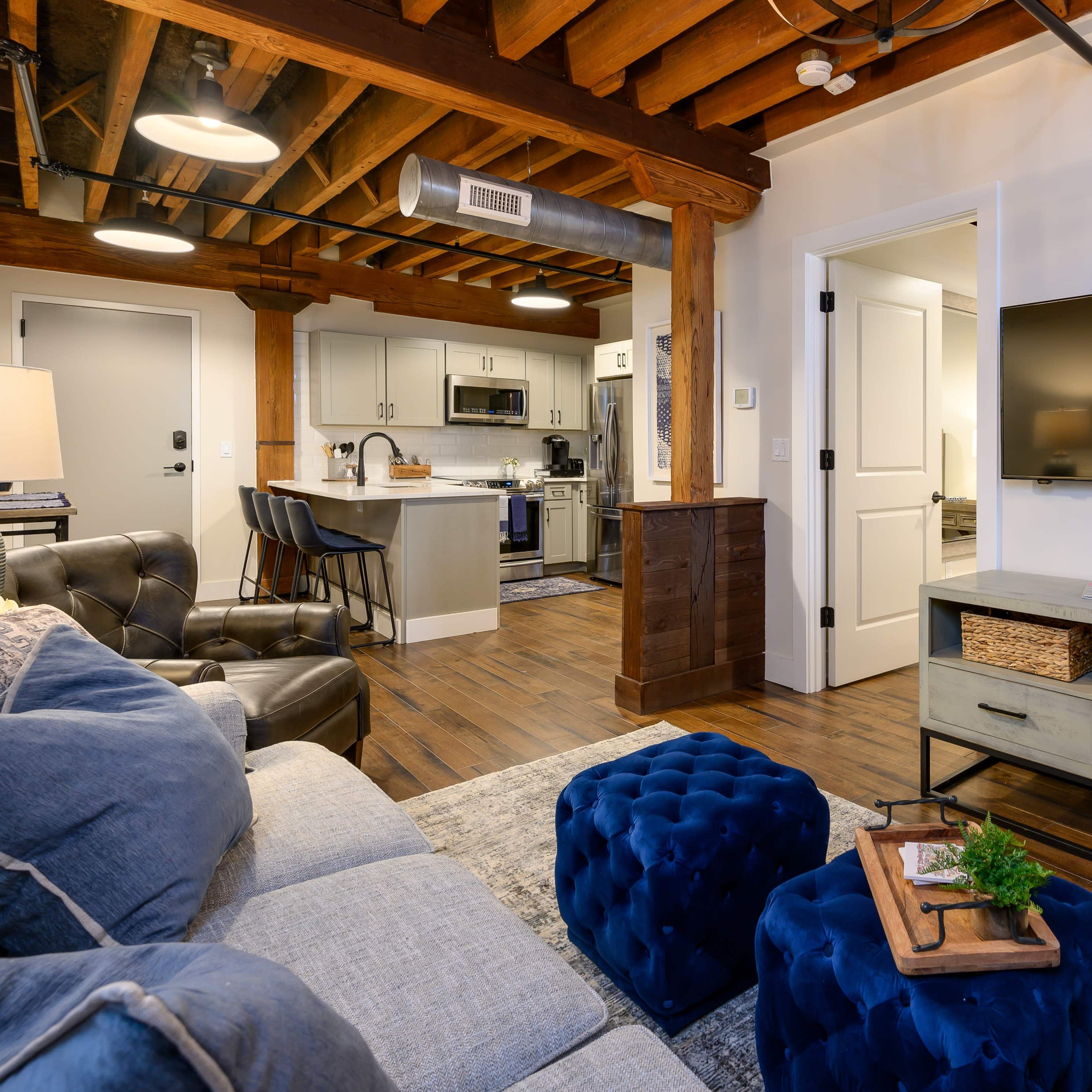 A modern living room with a cozy seating area featuring a gray sofa, blue ottomans, and a wall-mounted television, adjacent to a kitchen with white cabinets and a dining space.