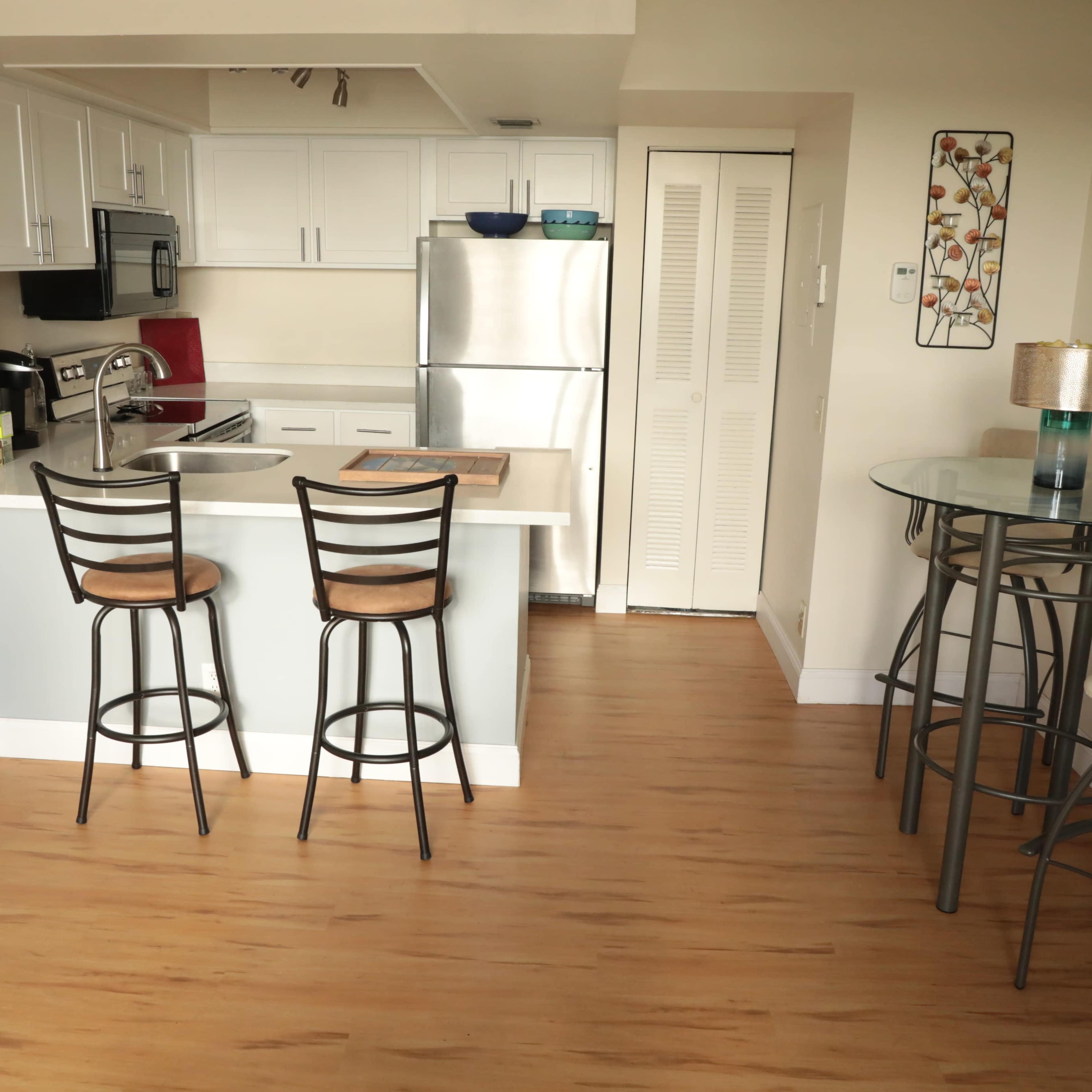A modern kitchen features white cabinets, stainless steel appliances, and a small dining area with two bar stools and a glass-topped table.
