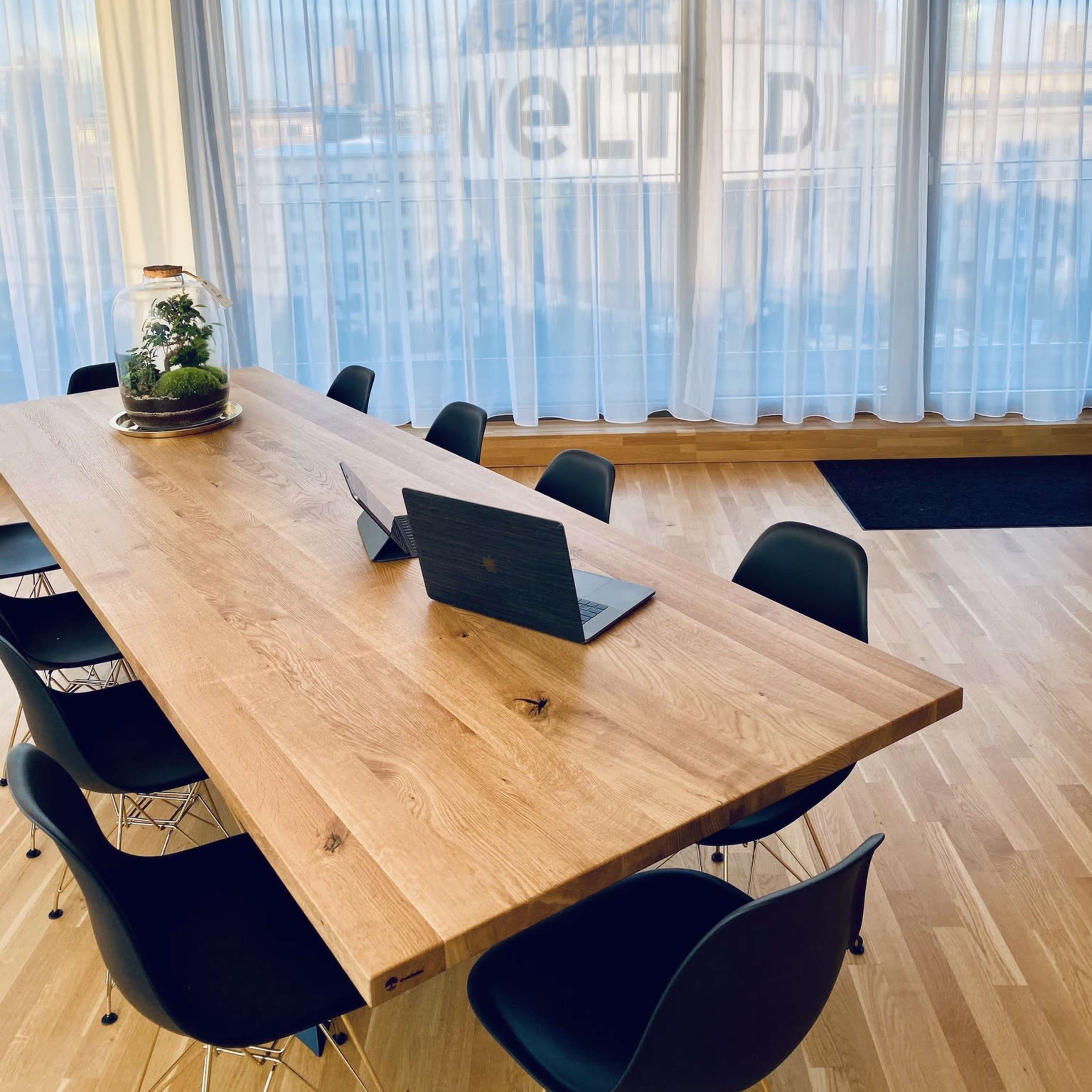 A modern conference room features a large wooden table with a laptop and chairs, illuminated by a geometric light fixture, and a view of a cityscape through sheer curtains.