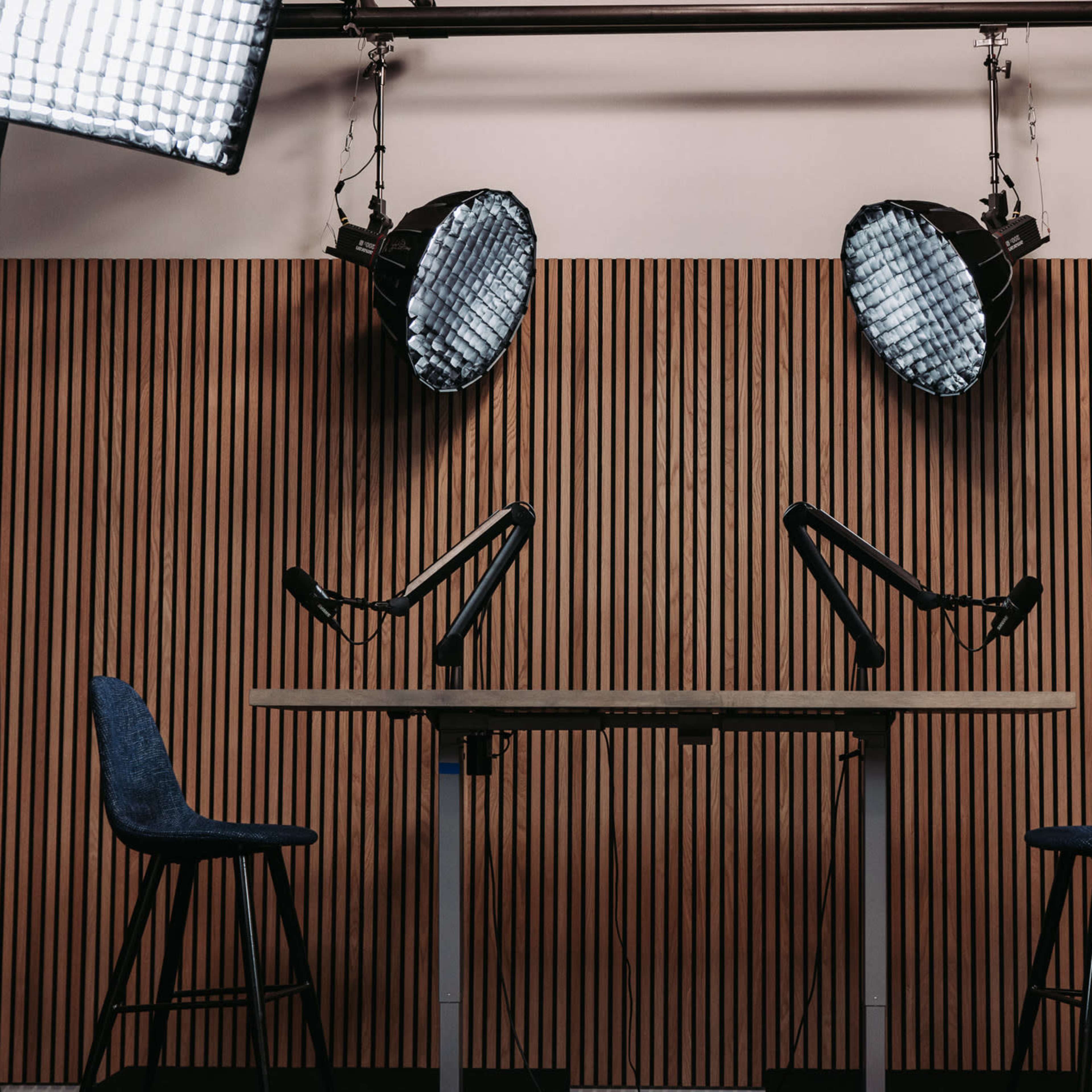 A podcast studio features a wooden wall, two microphone stands, and two chairs under softbox lights.