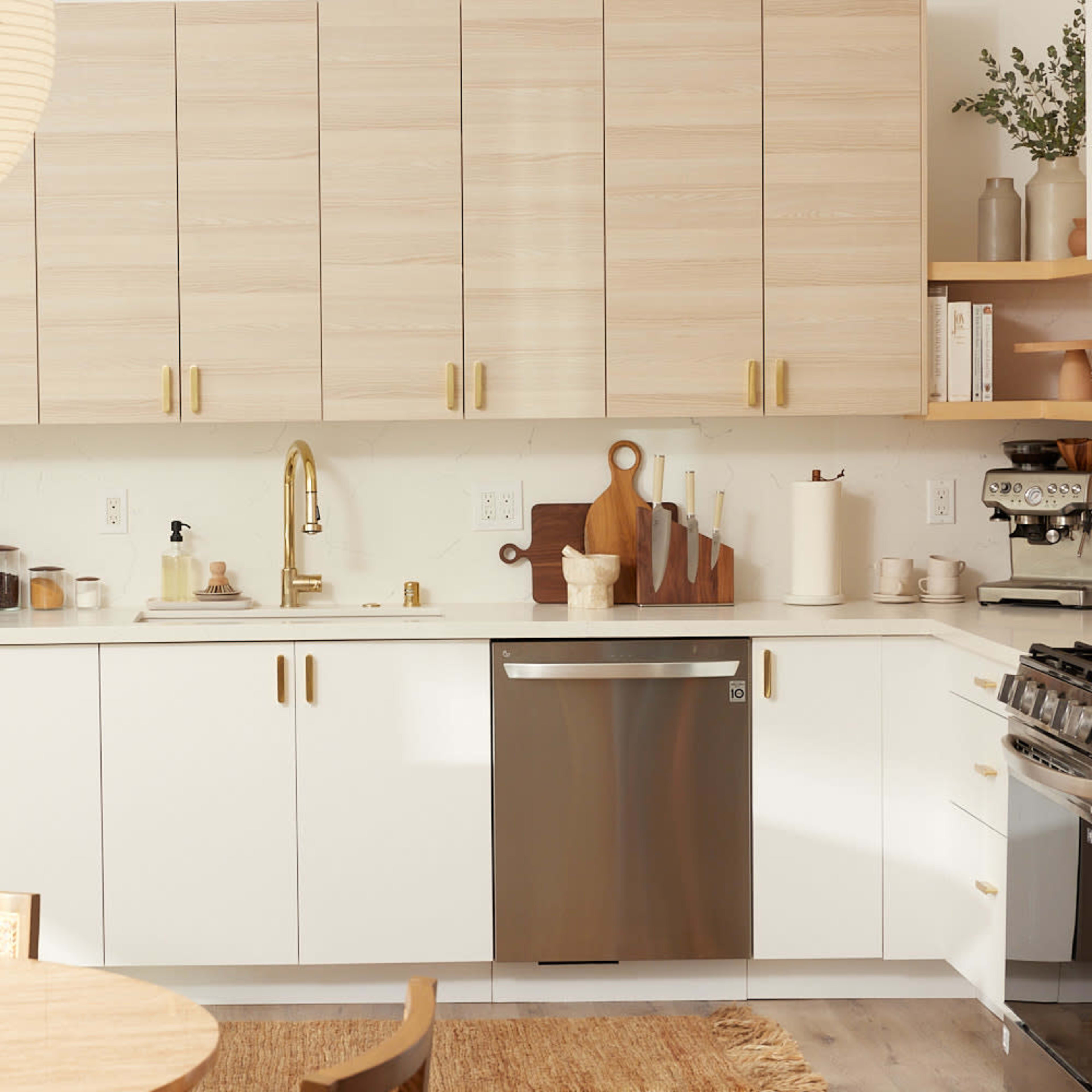 A modern kitchen with light wood cabinetry, a stainless steel dishwasher, and a round dining table with a centerpiece.