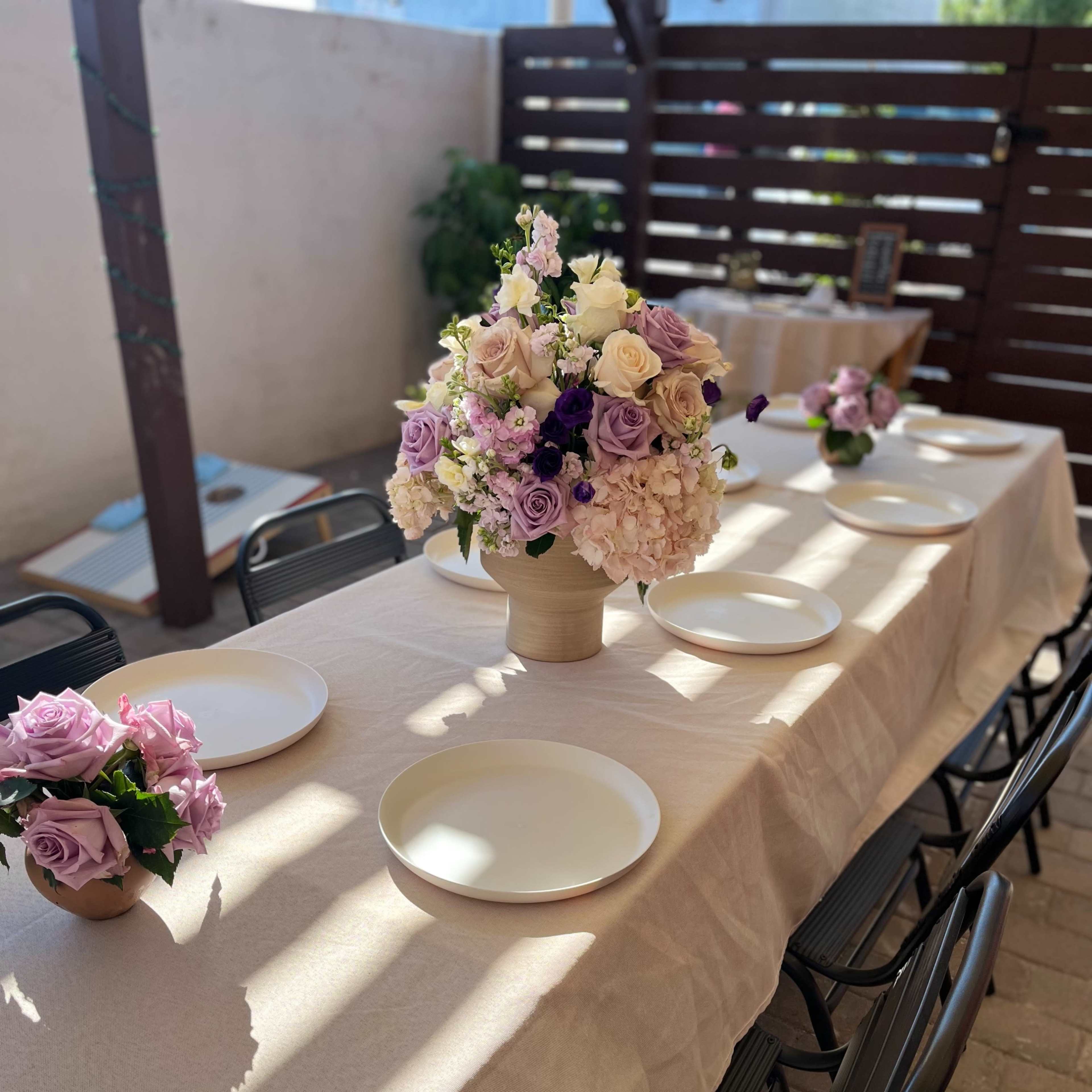 A long table is set with white plates and floral centerpieces in a well-lit outdoor space.