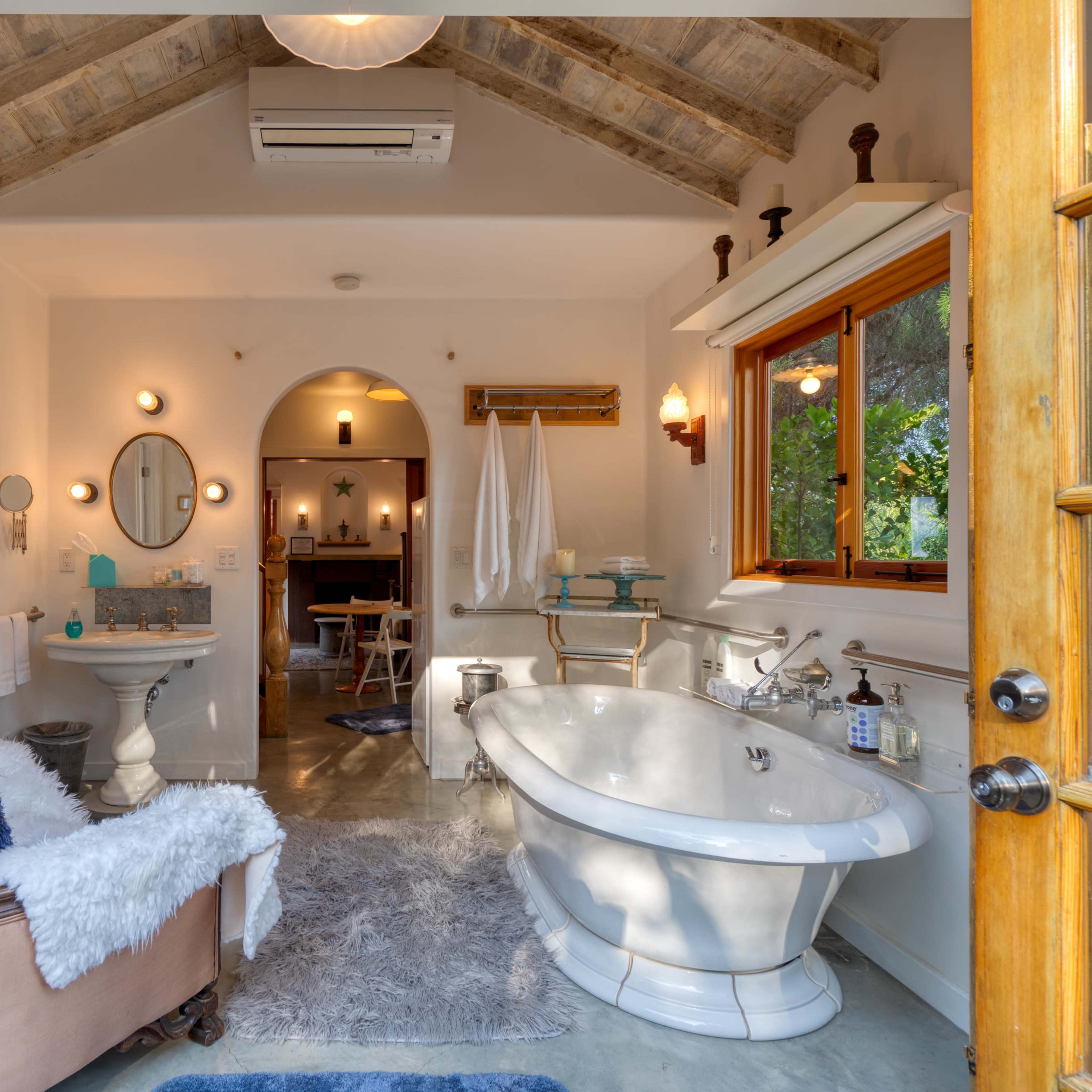 A spacious bathroom features a freestanding bathtub, a wooden armchair with a throw, and large windows allowing natural light.
