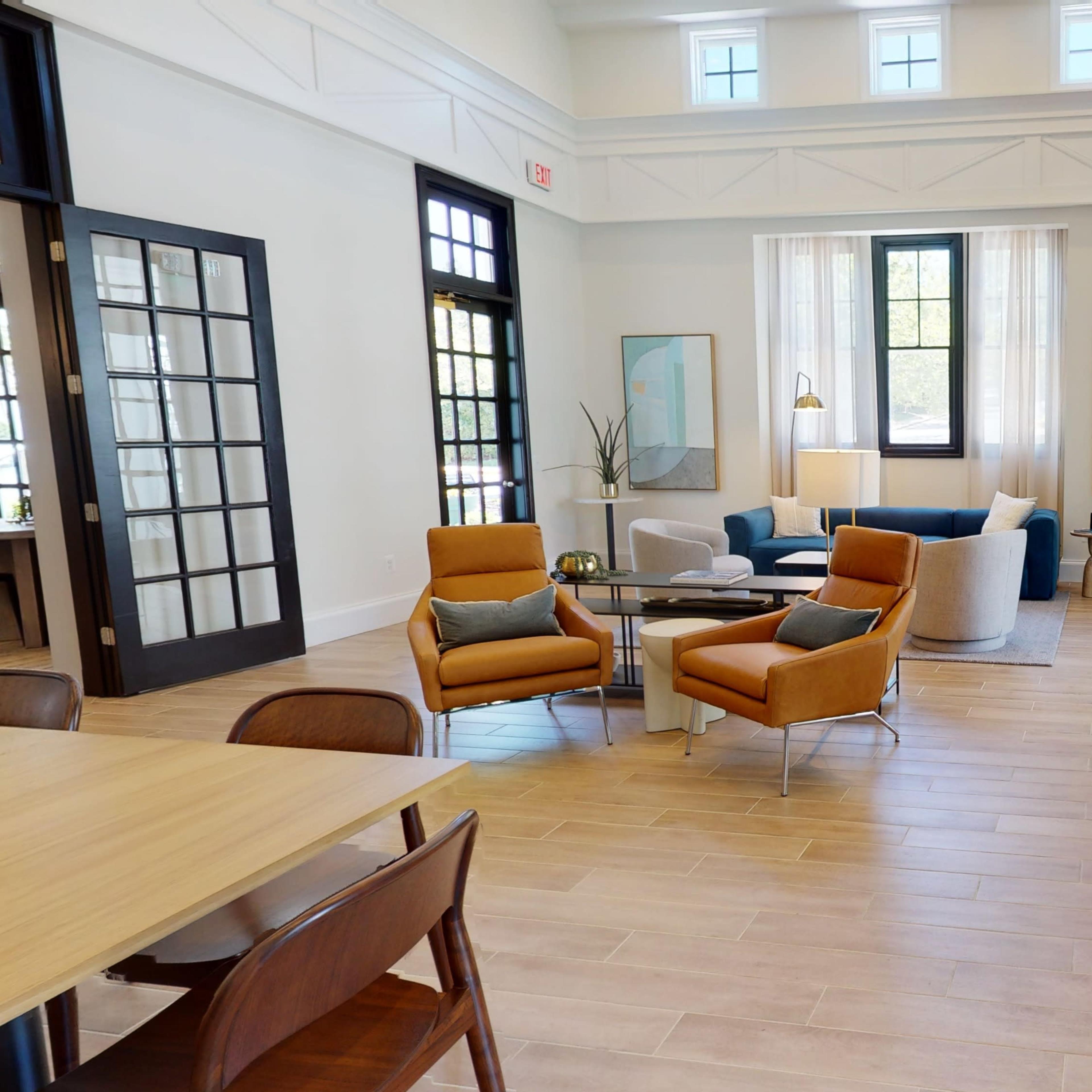 A bright, modern living area featuring a seating arrangement with orange chairs, a cream sofa, a coffee table, and a large fireplace, surrounded by tall windows and light-colored walls.