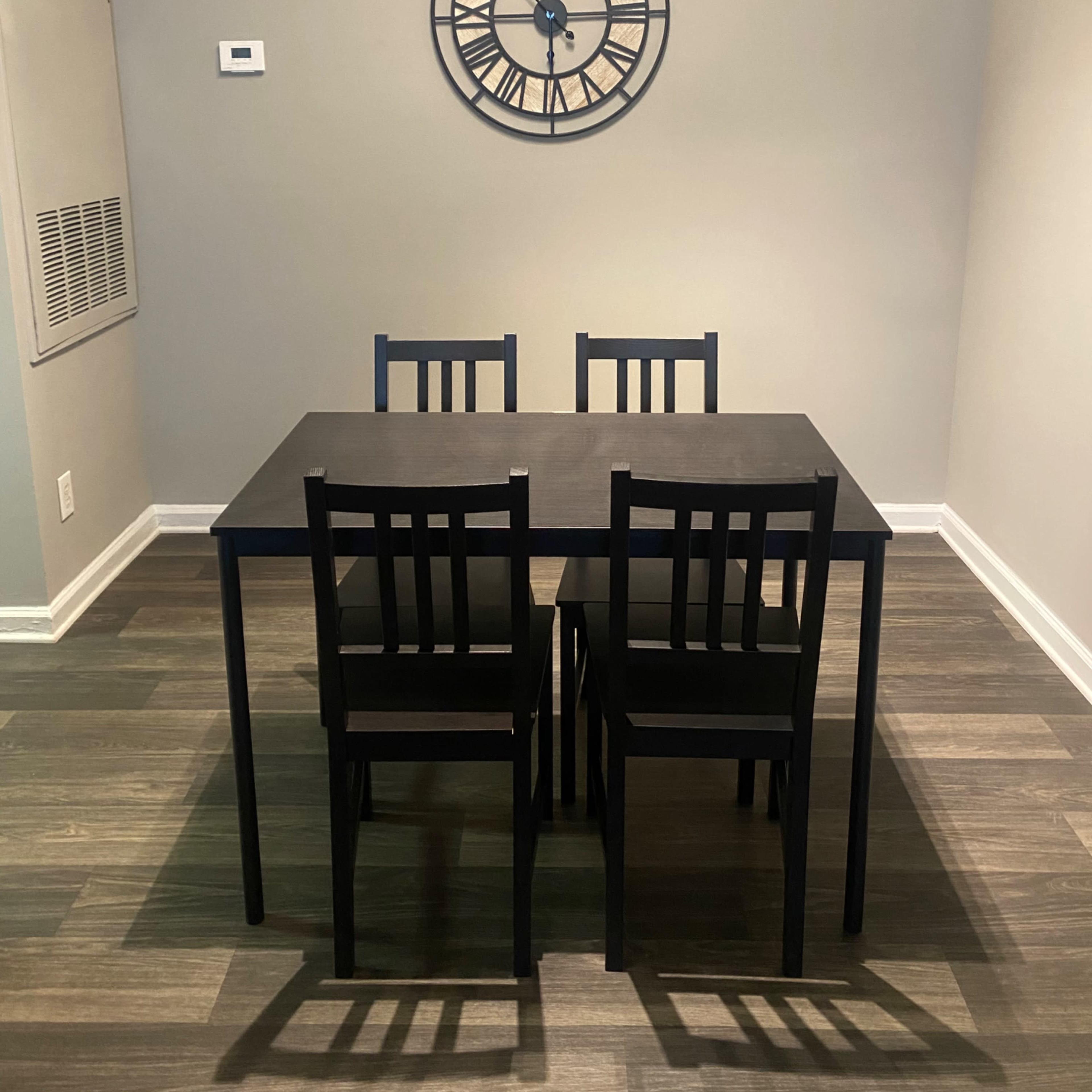 A black dining table with four chairs is positioned in a room featuring a clock on the wall and wooden flooring.