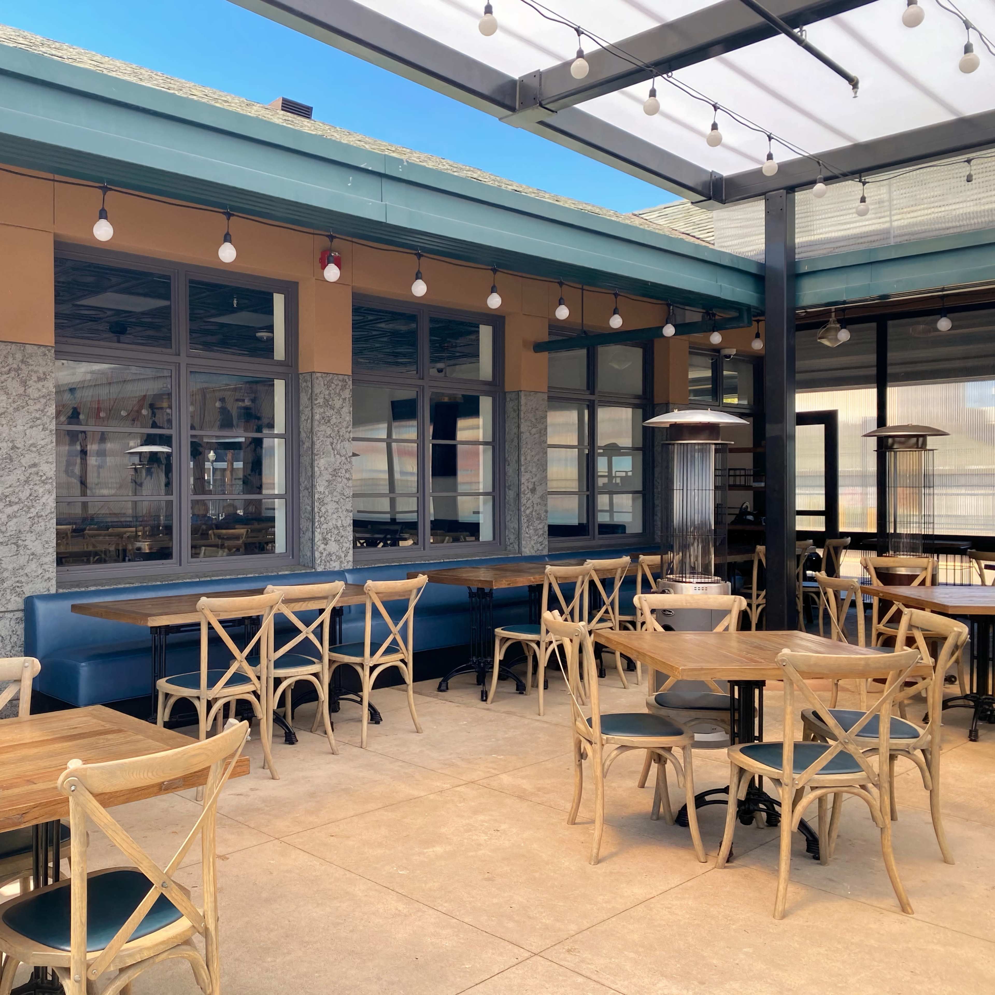 An outdoor dining area with wooden tables and chairs under a canopy, featuring string lights and a glass wall.