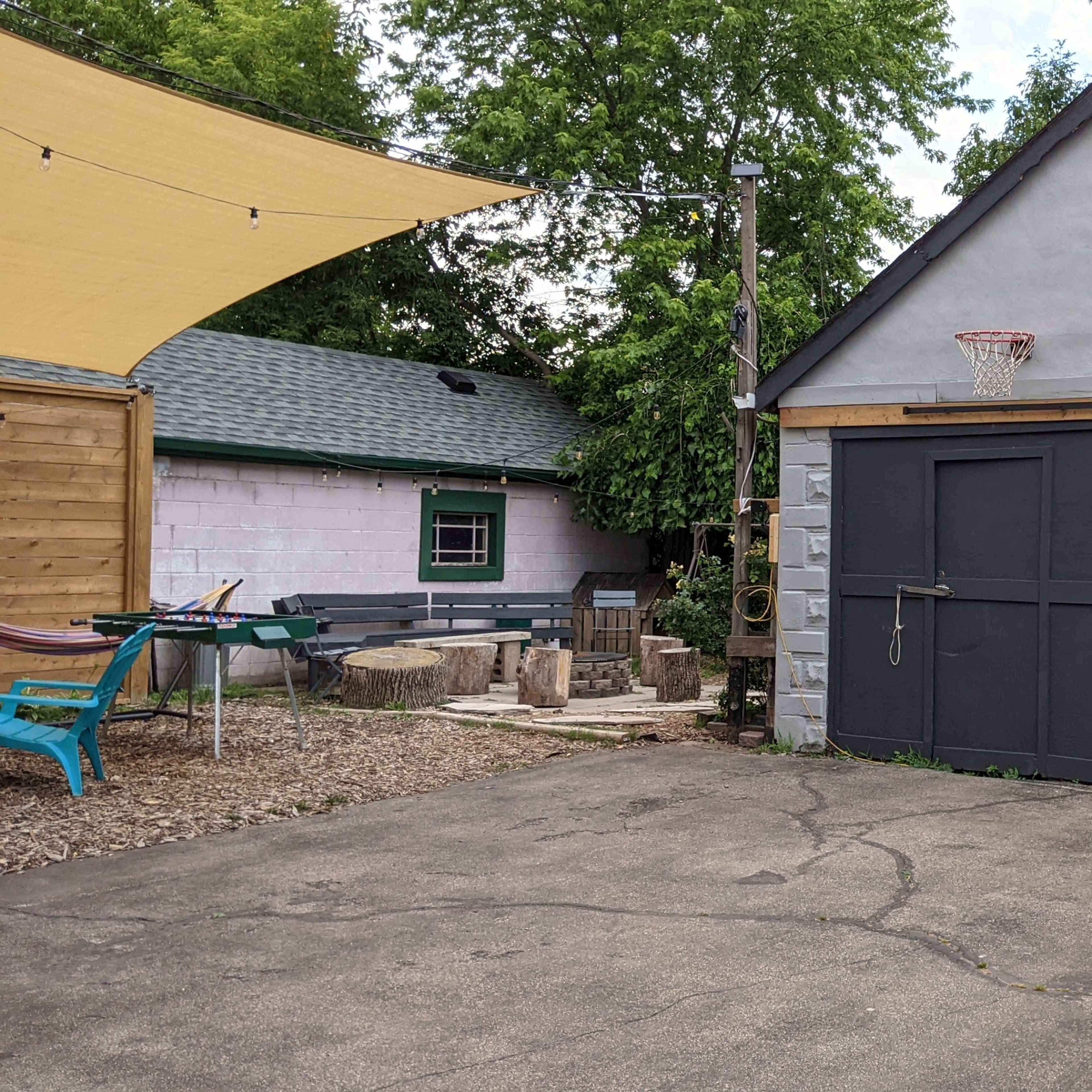 A garage with black double doors is situated next to a tree-lined yard featuring a basketball hoop and various outdoor furniture.