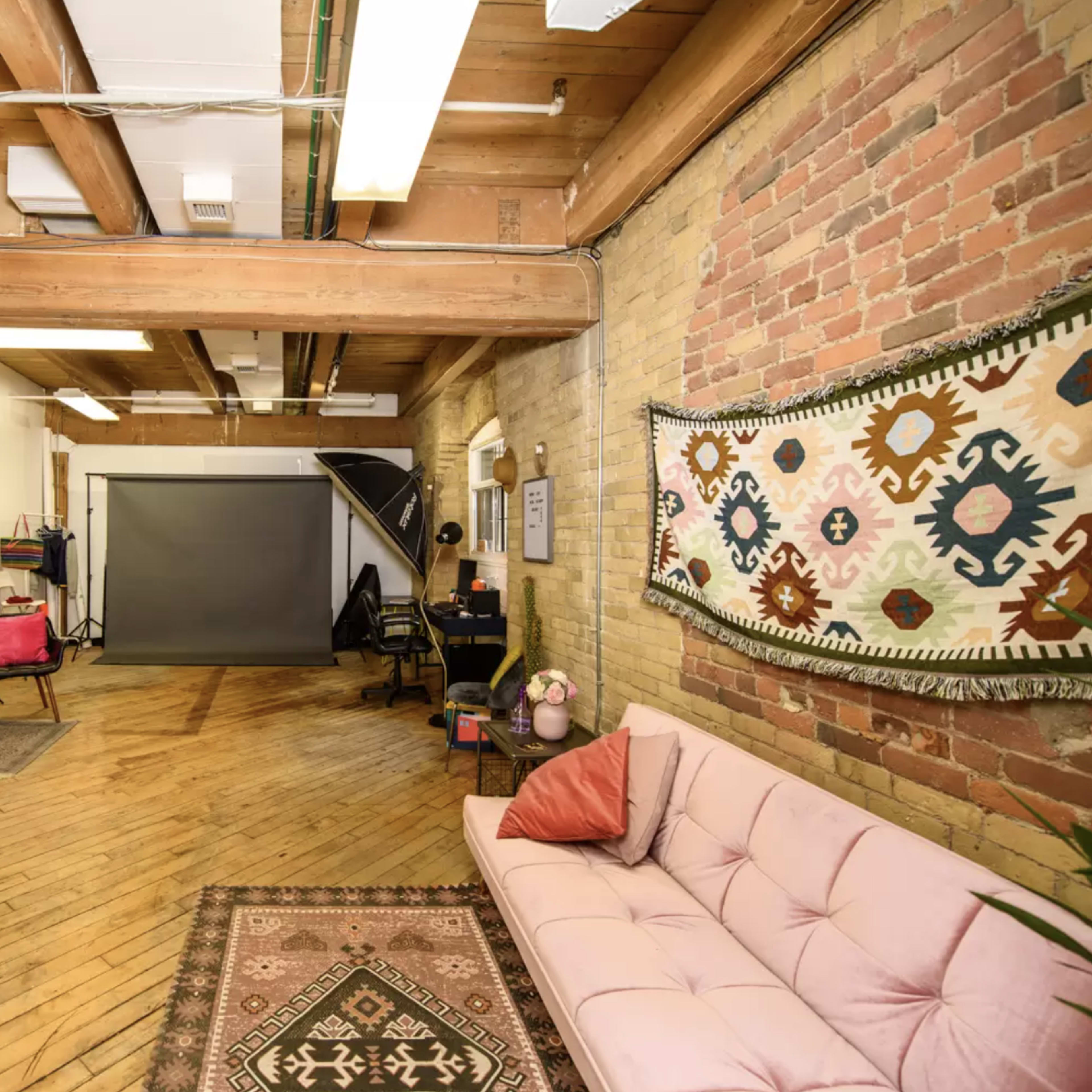 A spacious studio with wooden beams, brick walls, and a pink couch, featuring a backdrop for photography and various furniture arranged around the room.