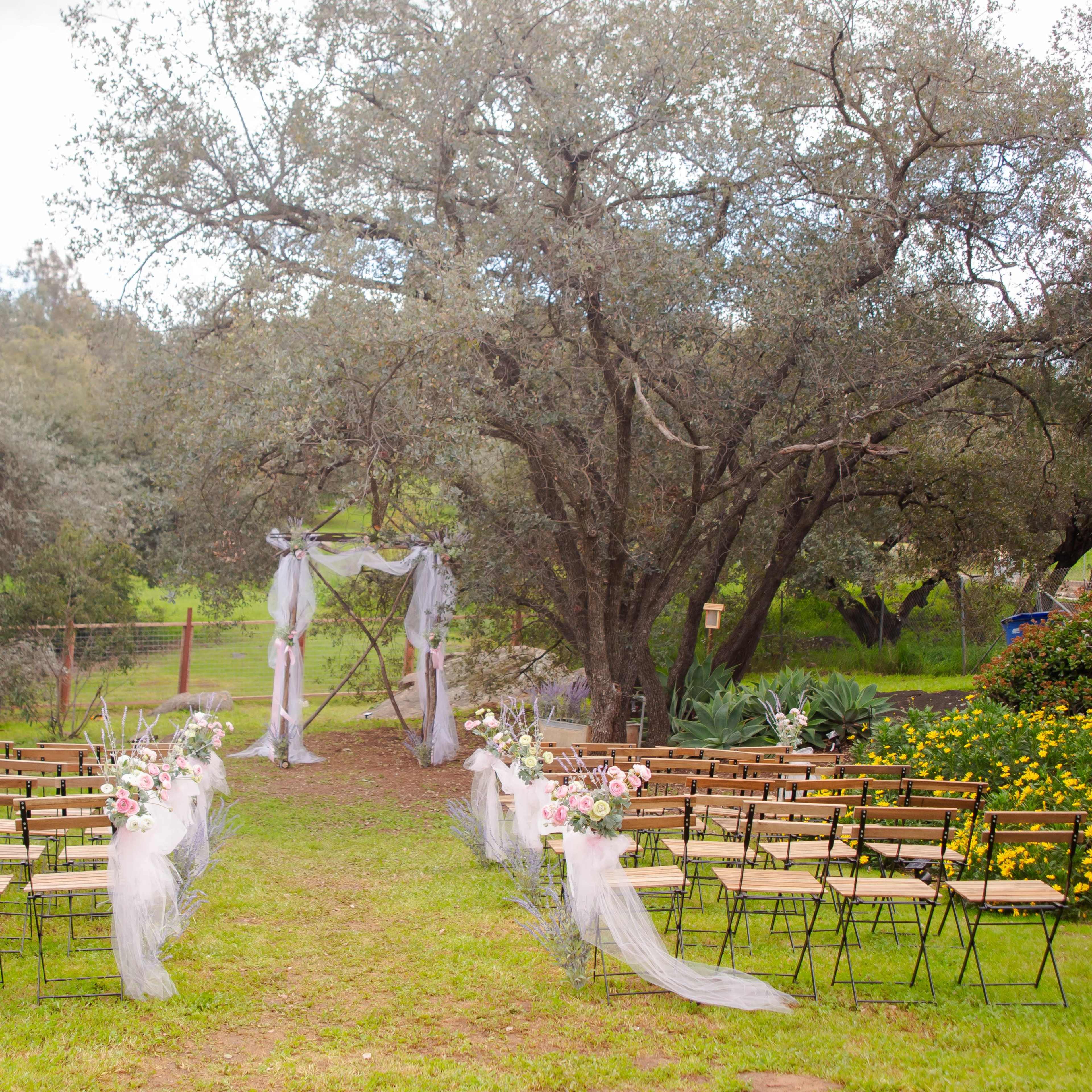 A wedding ceremony setup features rows of chairs arranged on a grassy area beneath large trees, with floral decorations and draped fabric framing an altar.
