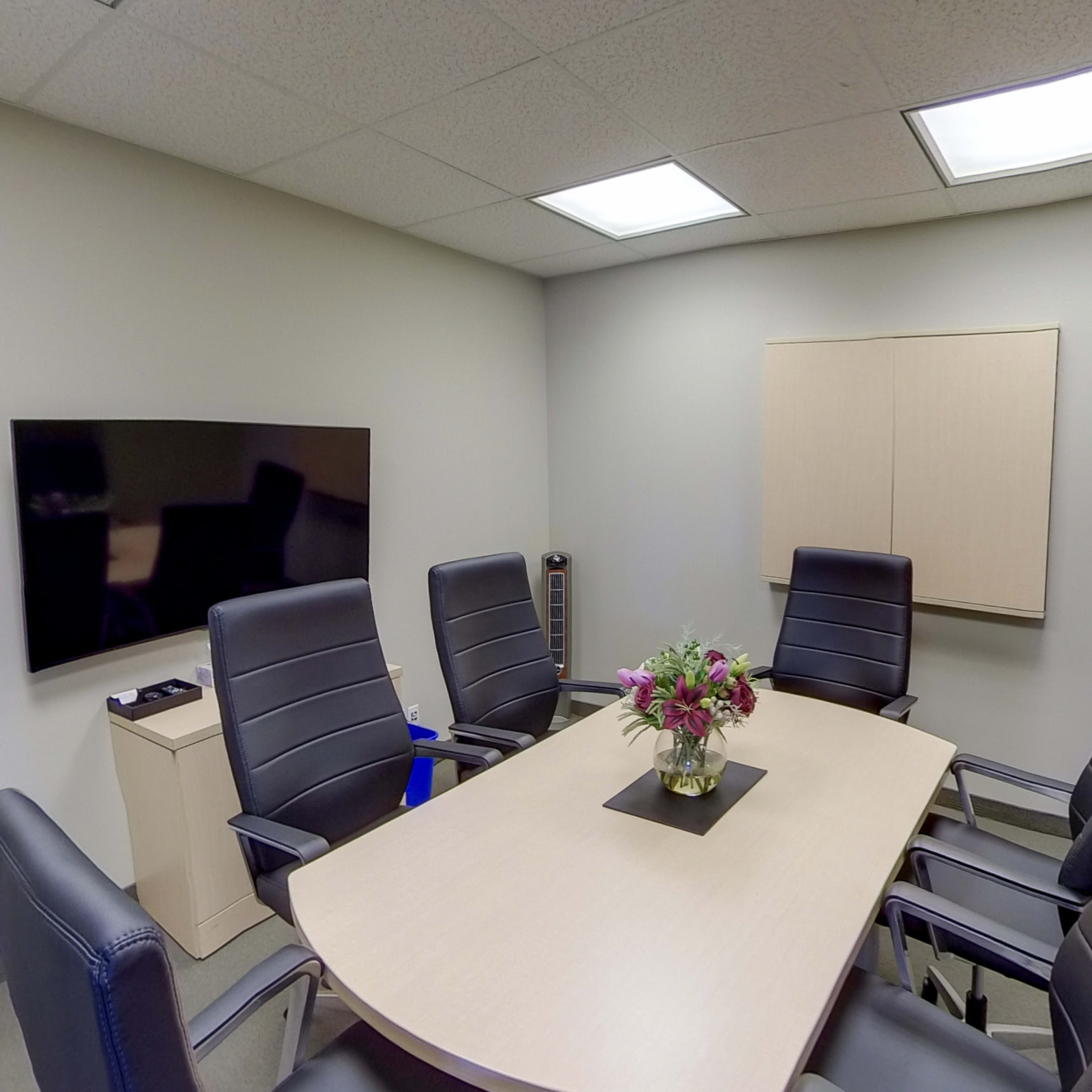 A conference room features a large table surrounded by six black chairs, with a television on the wall and a vase of flowers in the center.