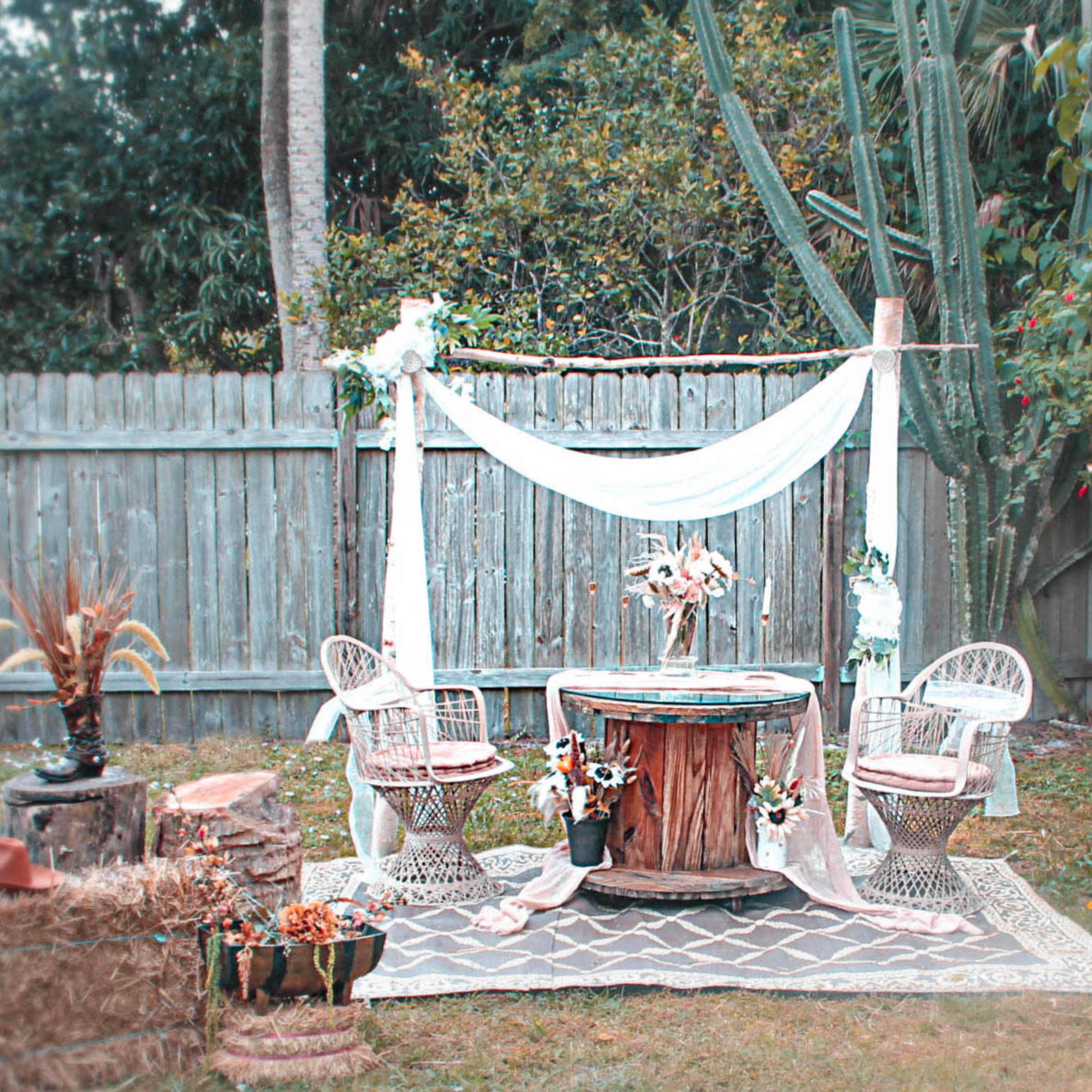 A backyard space is set up with a wooden table, two wicker chairs, and decorative elements surrounded by straw bales and plants.
