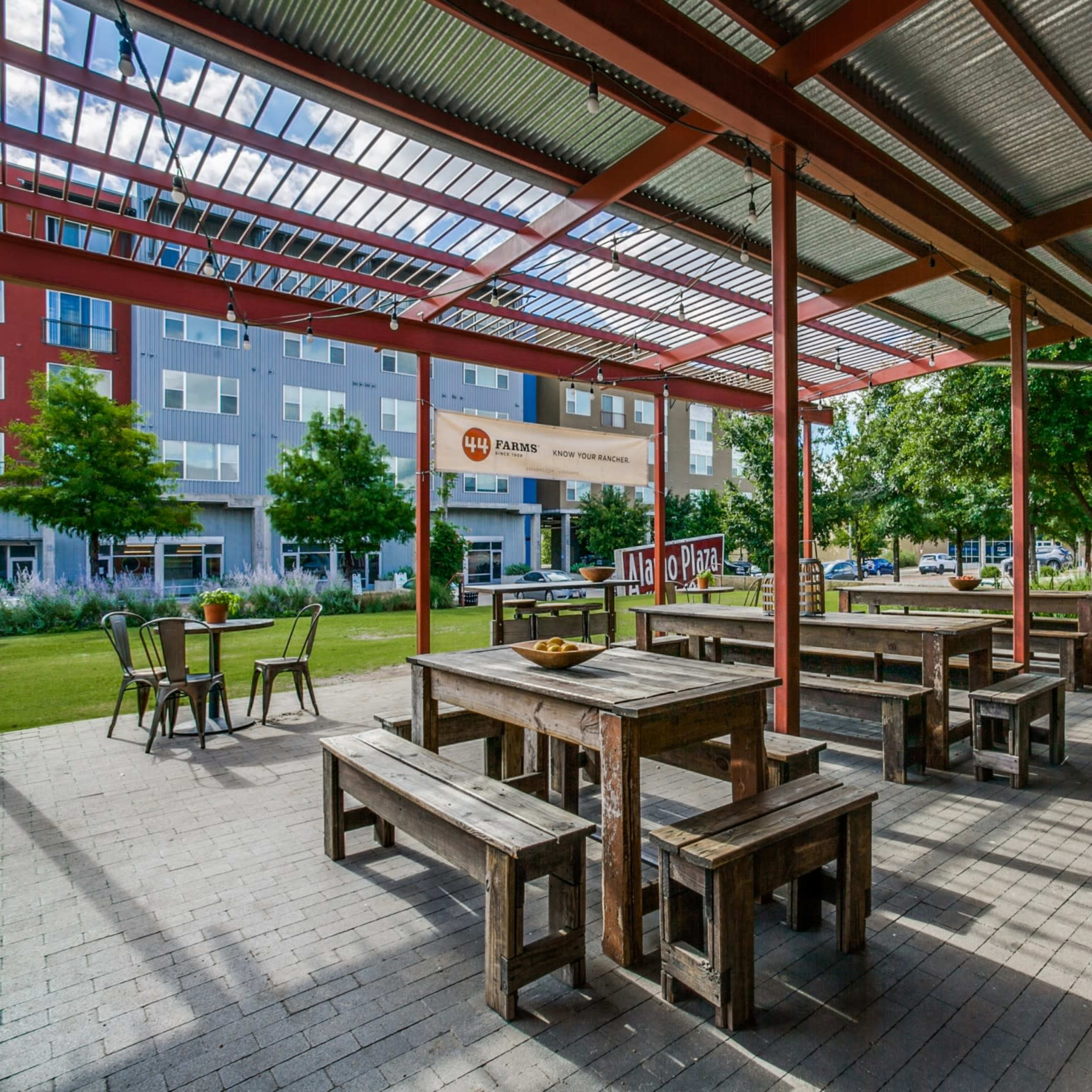 An outdoor seating area with wooden benches and tables under a metal roof, surrounded by greenery and modern buildings.