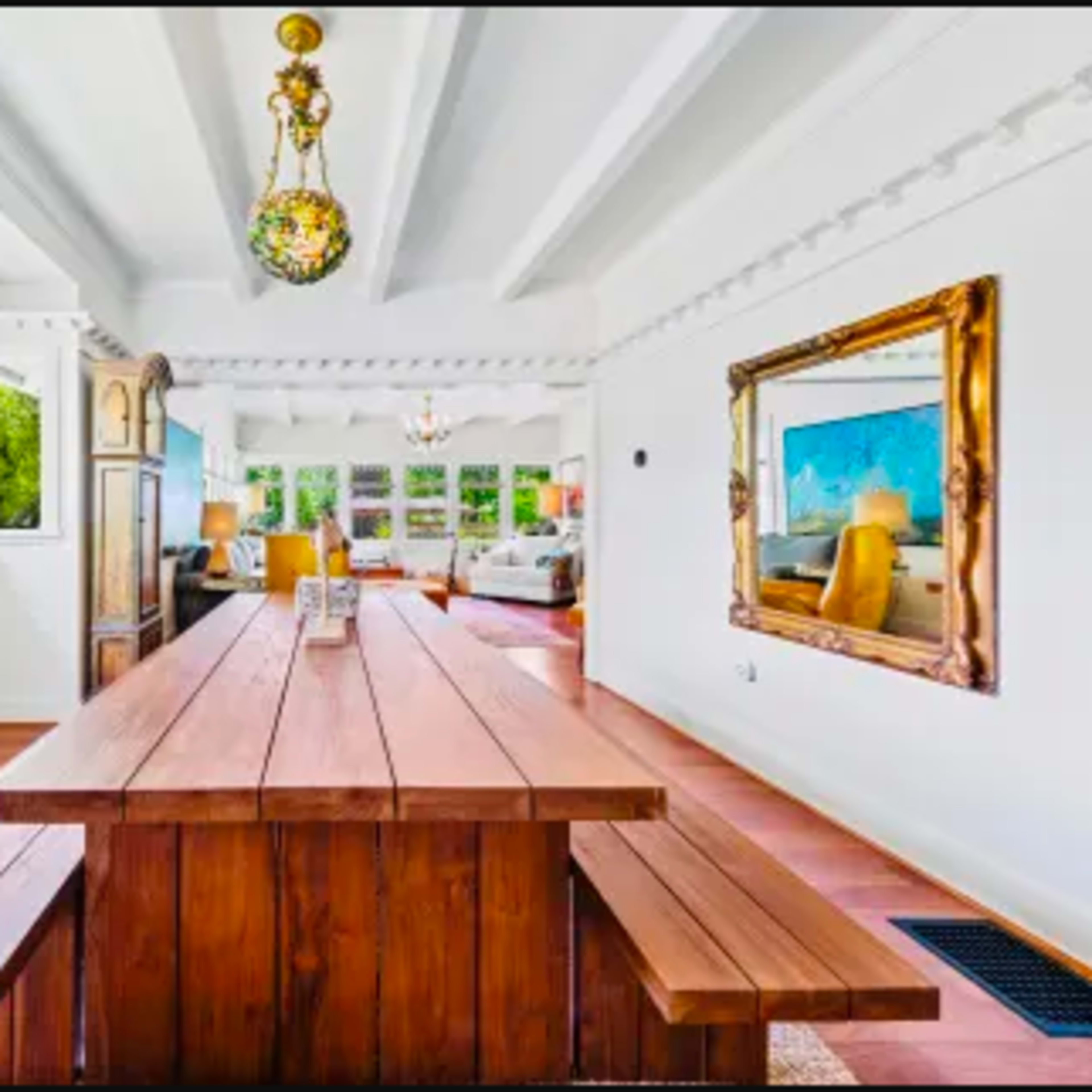A bright, open dining area with a long wooden table and benches, featuring large windows that look out onto greenery.