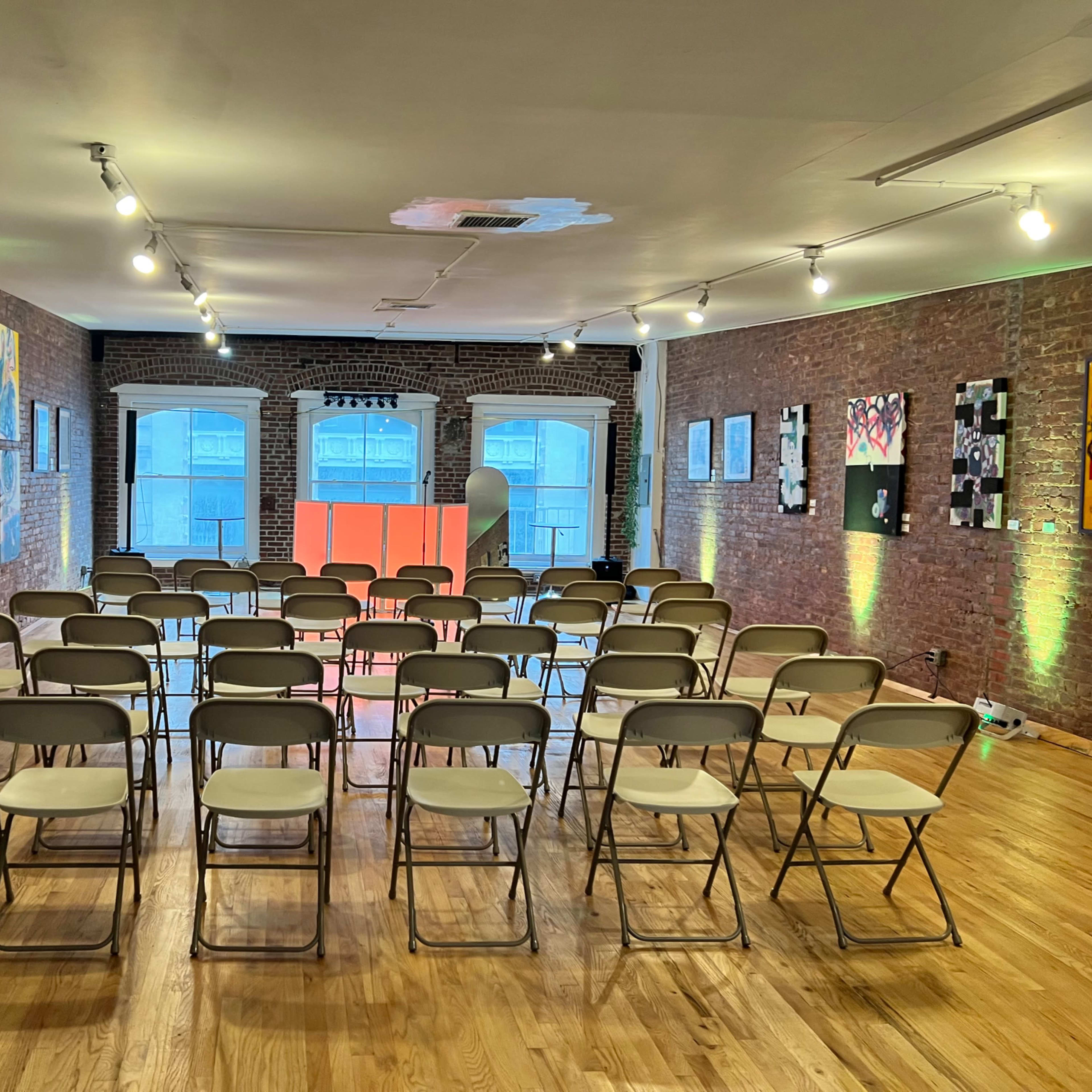 A room set up with rows of folding chairs facing a small stage, with exposed brick walls and various pieces of artwork displayed.