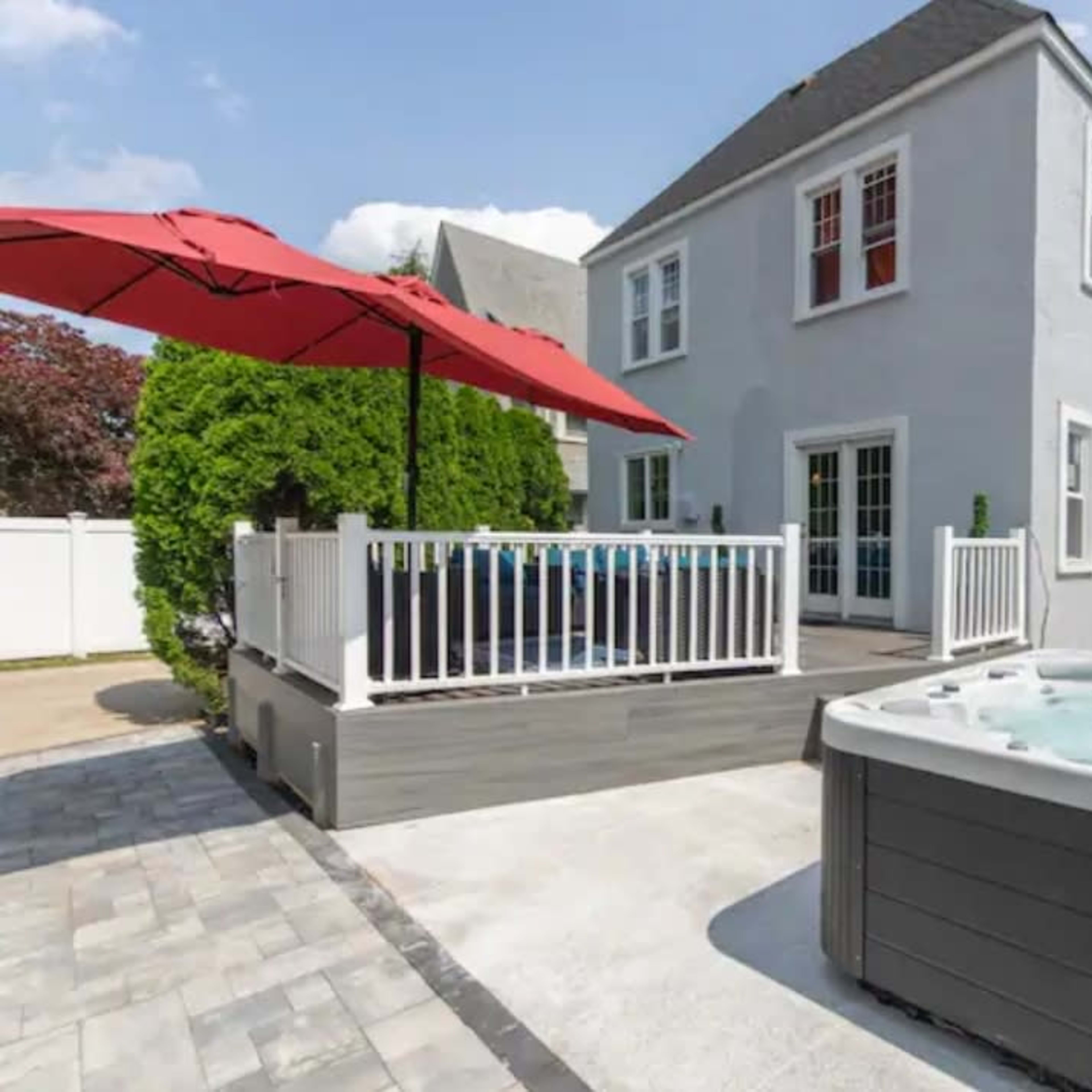 A backyard with a hot tub adjacent to a deck featuring seating under a red umbrella, beside a two-story gray house.